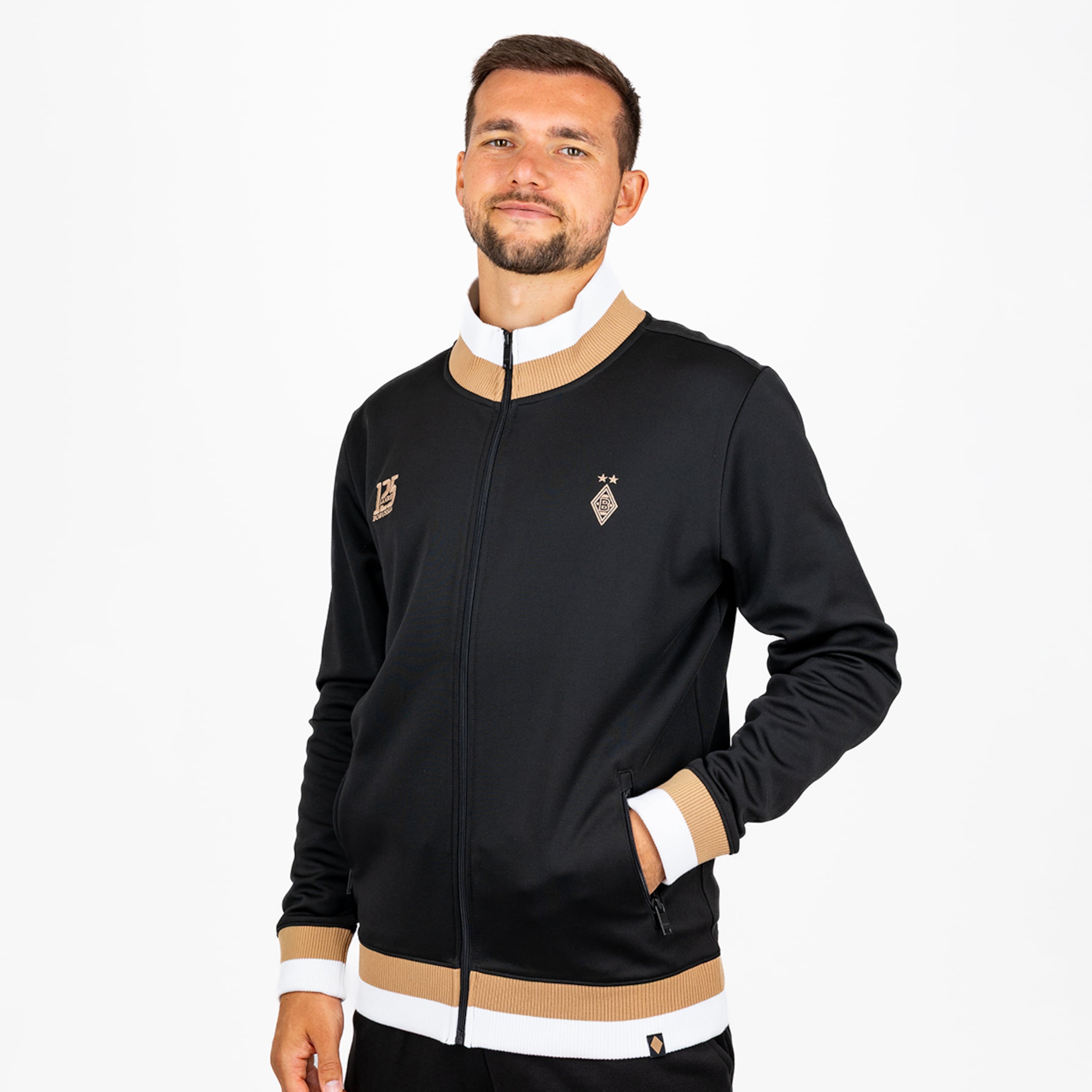 Mann in schwarzer Borussia Mönchengladbach 125-Jahre-Jubiläumsjacke mit gold-weißen Details und Vereinslogo vor weißem Hintergrund.