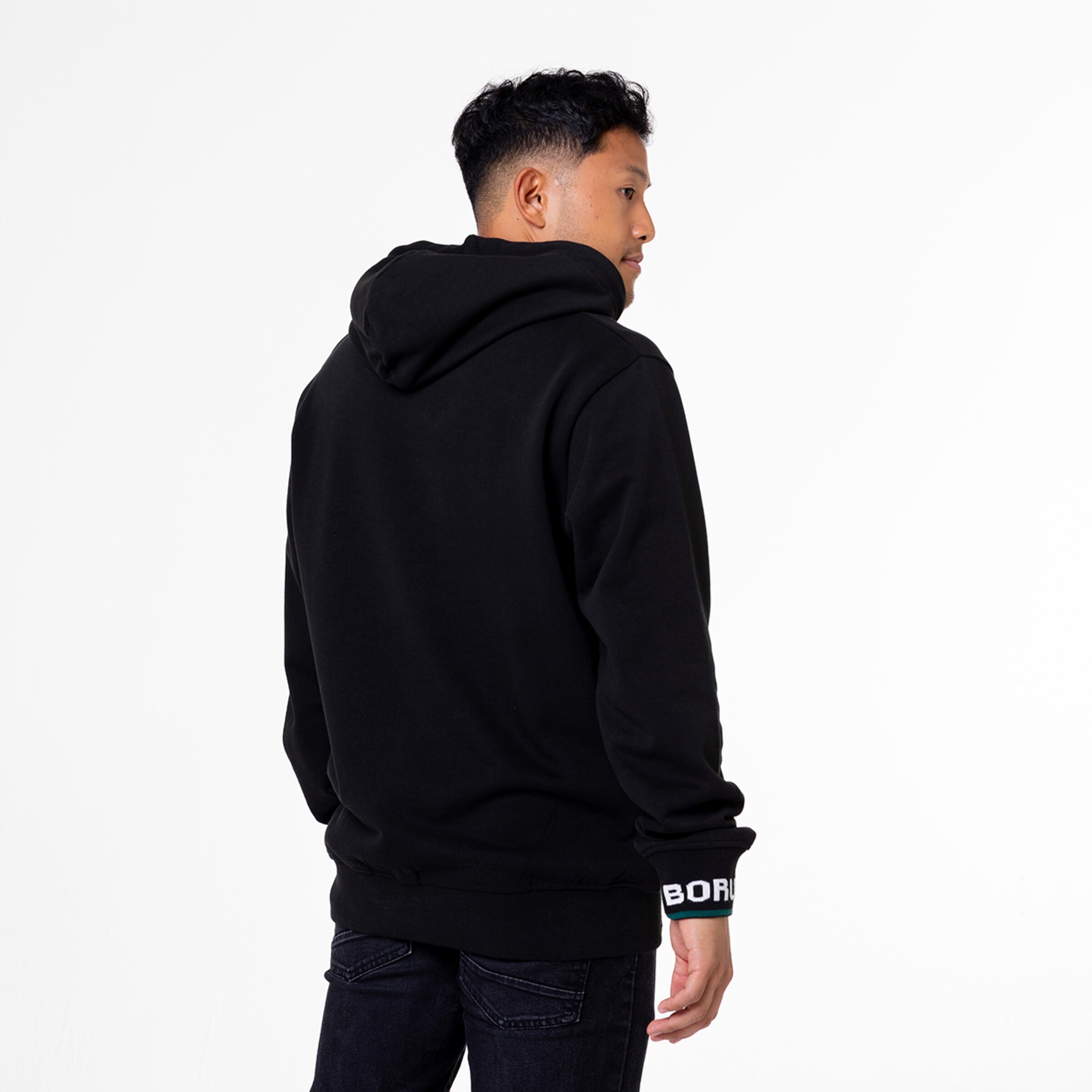 Man in black hoodie and black jeans viewed from the back standing against a white background.