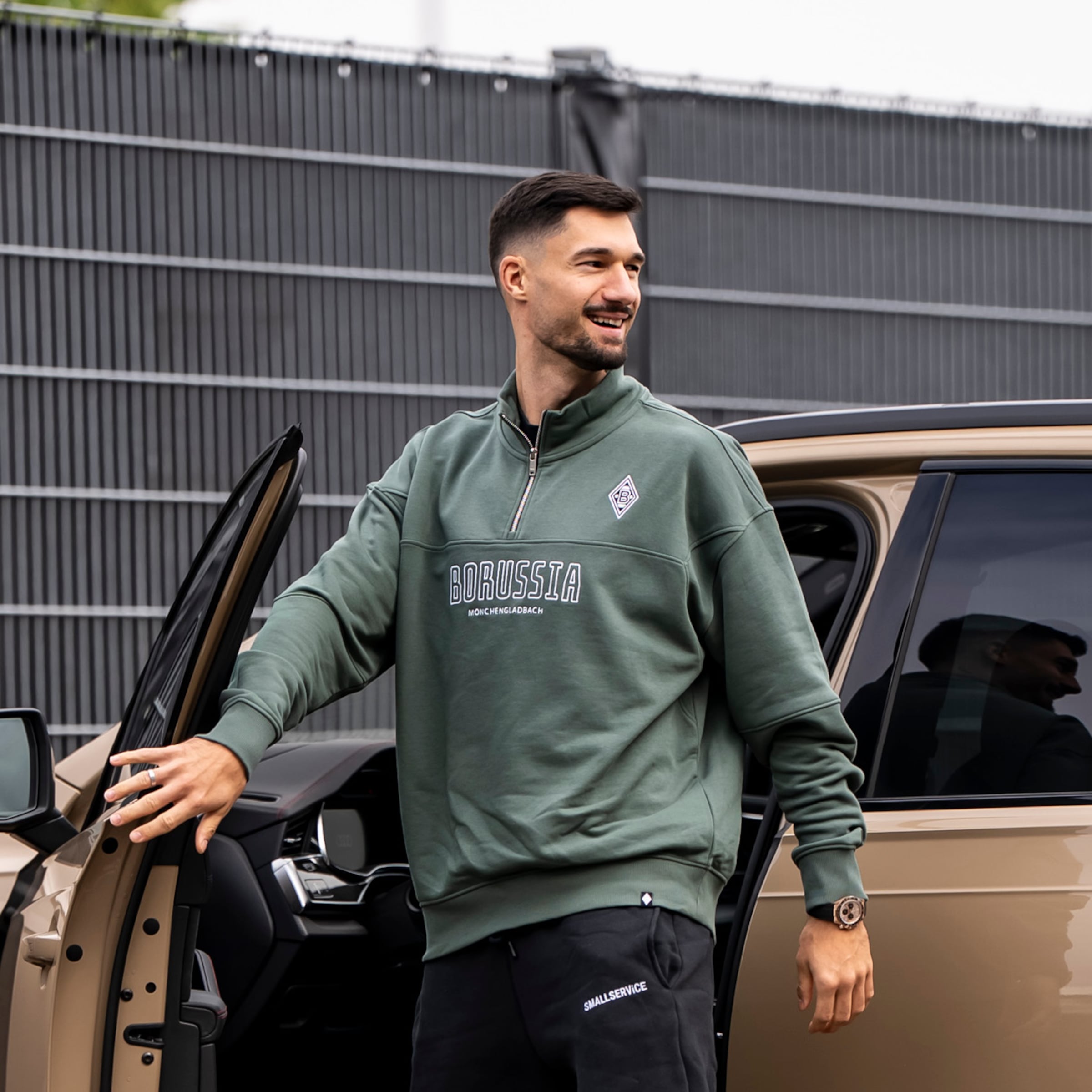 Lächelnder Mann in grünem Borussia Mönchengladbach Sweatshirt steht an einer offenen Autotür vor einem grauen Zaun.