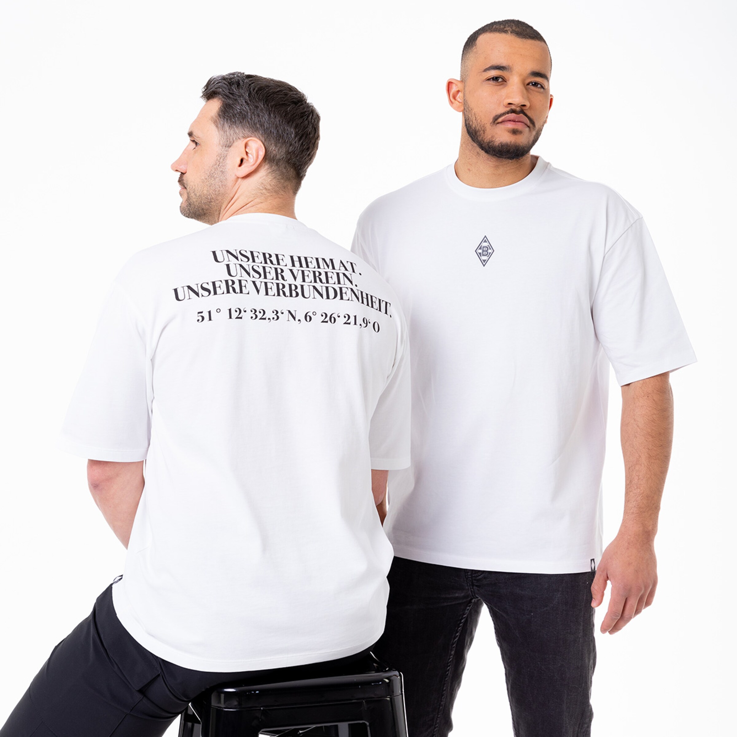 Zwei Männer tragen weiße T-Shirts mit einem Schriftzug auf dem Rücken: UNSERE HEIMAT. UNSER VEREIN. UNSERE VERBUNDENHEIT. und Koordinaten.