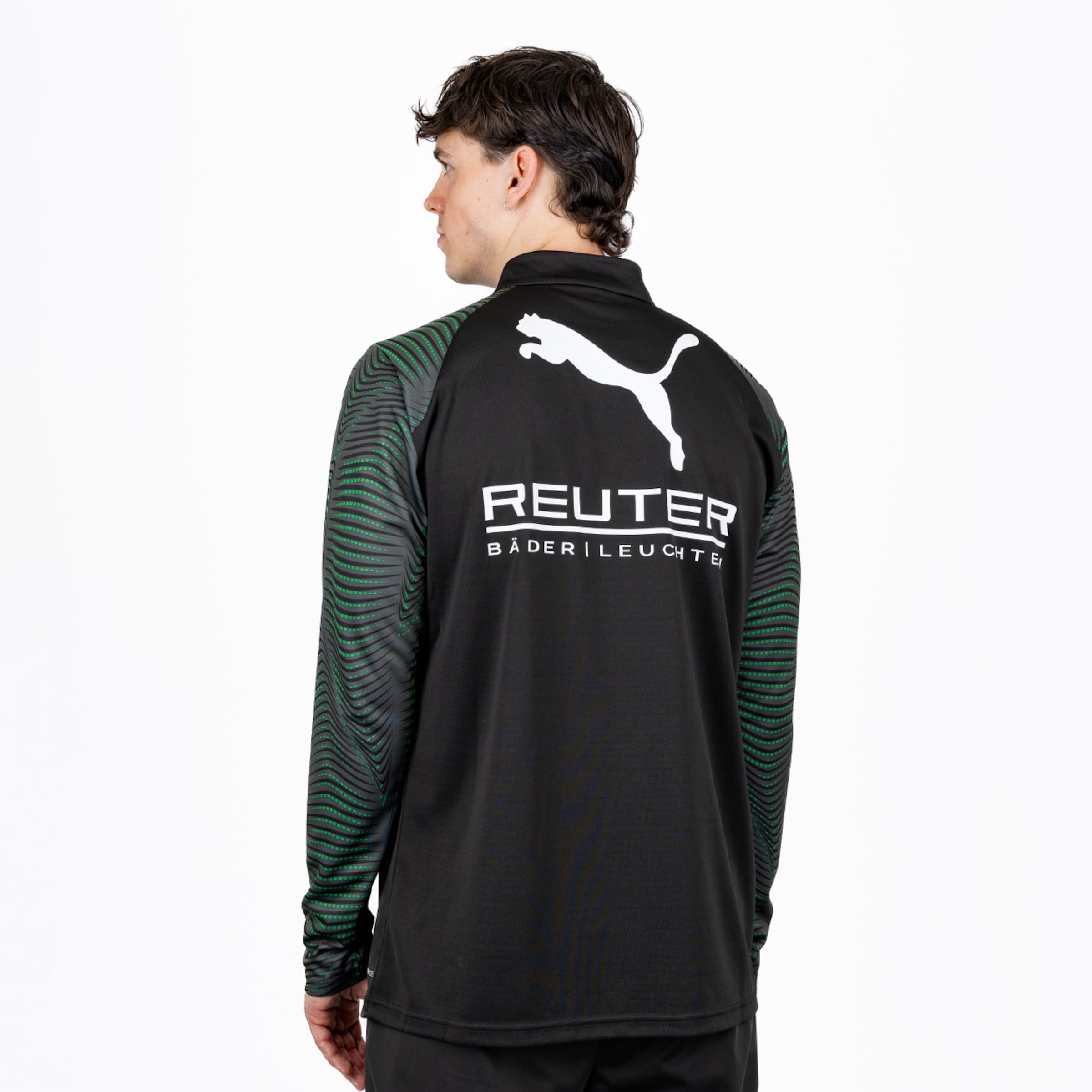Rückenansicht eines Mannes in einem schwarzen Puma Langarmshirt mit grünen Wellenmustern auf den Ärmeln und dem Schriftzug 'REUTER BÄDERLEUCHTE'. Er blickt nach links.