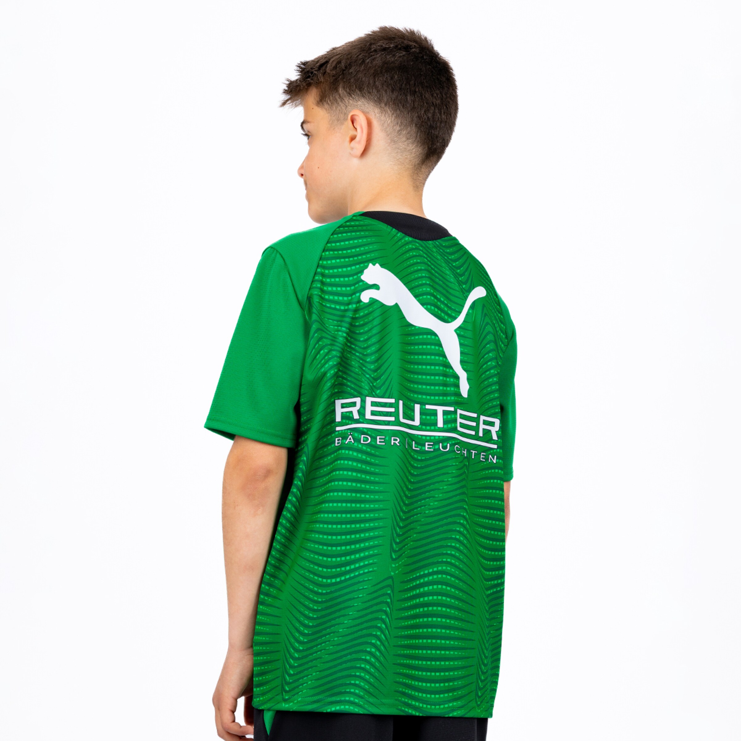 Rückenansicht eines Jungen in einem grünen Puma-Fußballtrikot mit strukturiertem Muster. Das Trikot zeigt ein weißes Puma-Logo und den Schriftzug „REUTER BÄDER LEUCHTEN“ auf dem Rücken vor weißem Hintergrund.