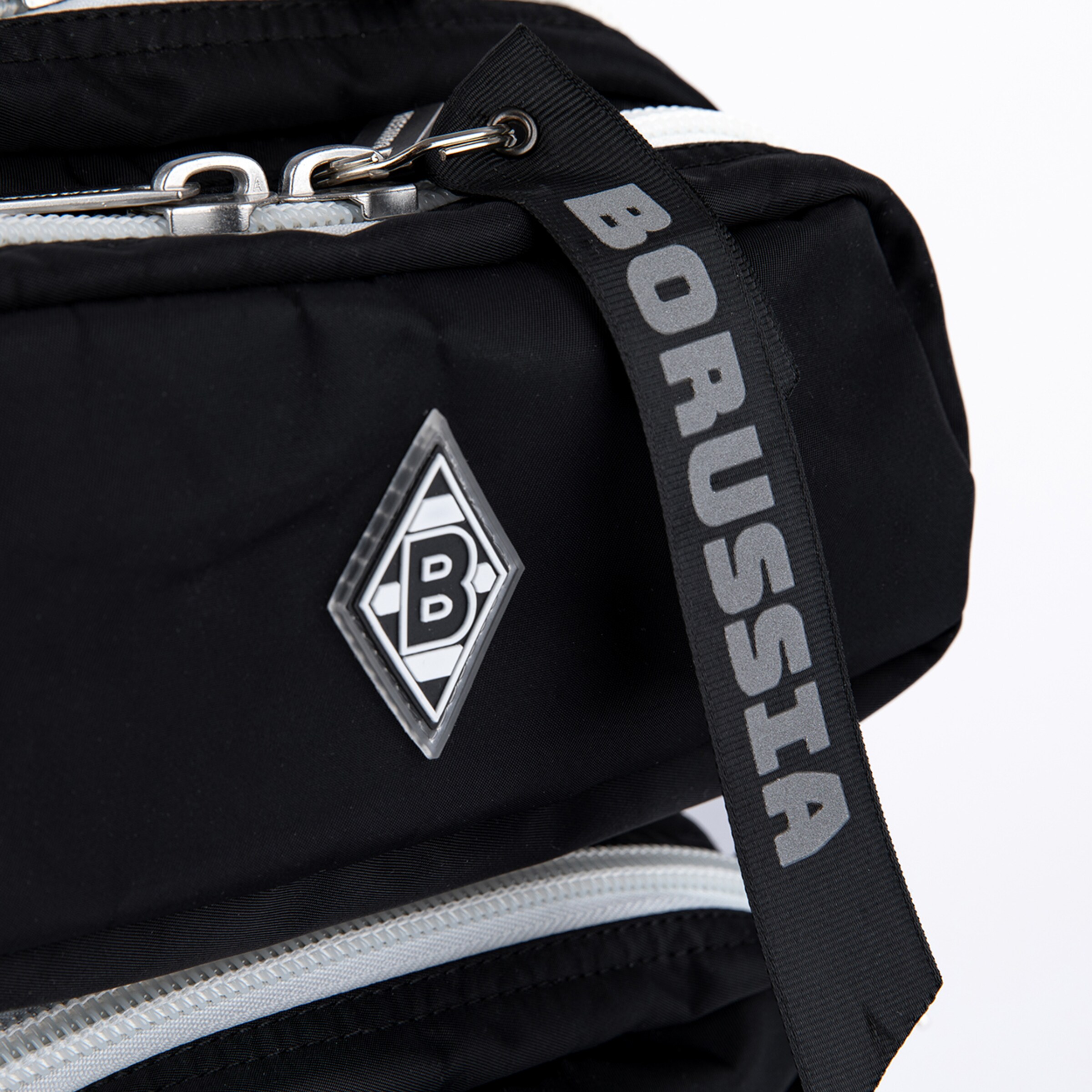 Close-up of a black bag with a white zipper, featuring the Borussia Mönchengladbach logo—a white 'B' inside a diamond shape. A black strap with 'BORUSSIA' in grey lettering is attached to the bag.