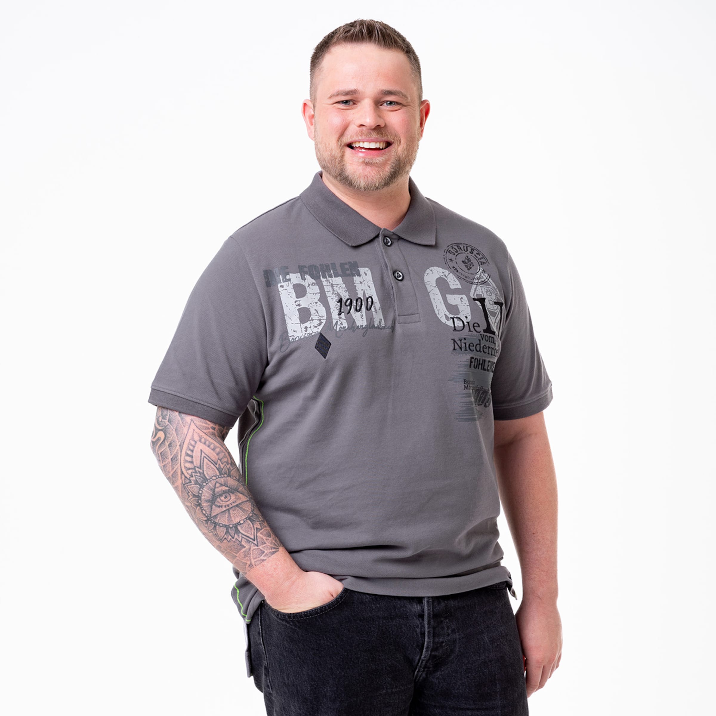 Lächelnder Mann mit Tätowierung trägt graues Poloshirt und schwarze Jeans.
