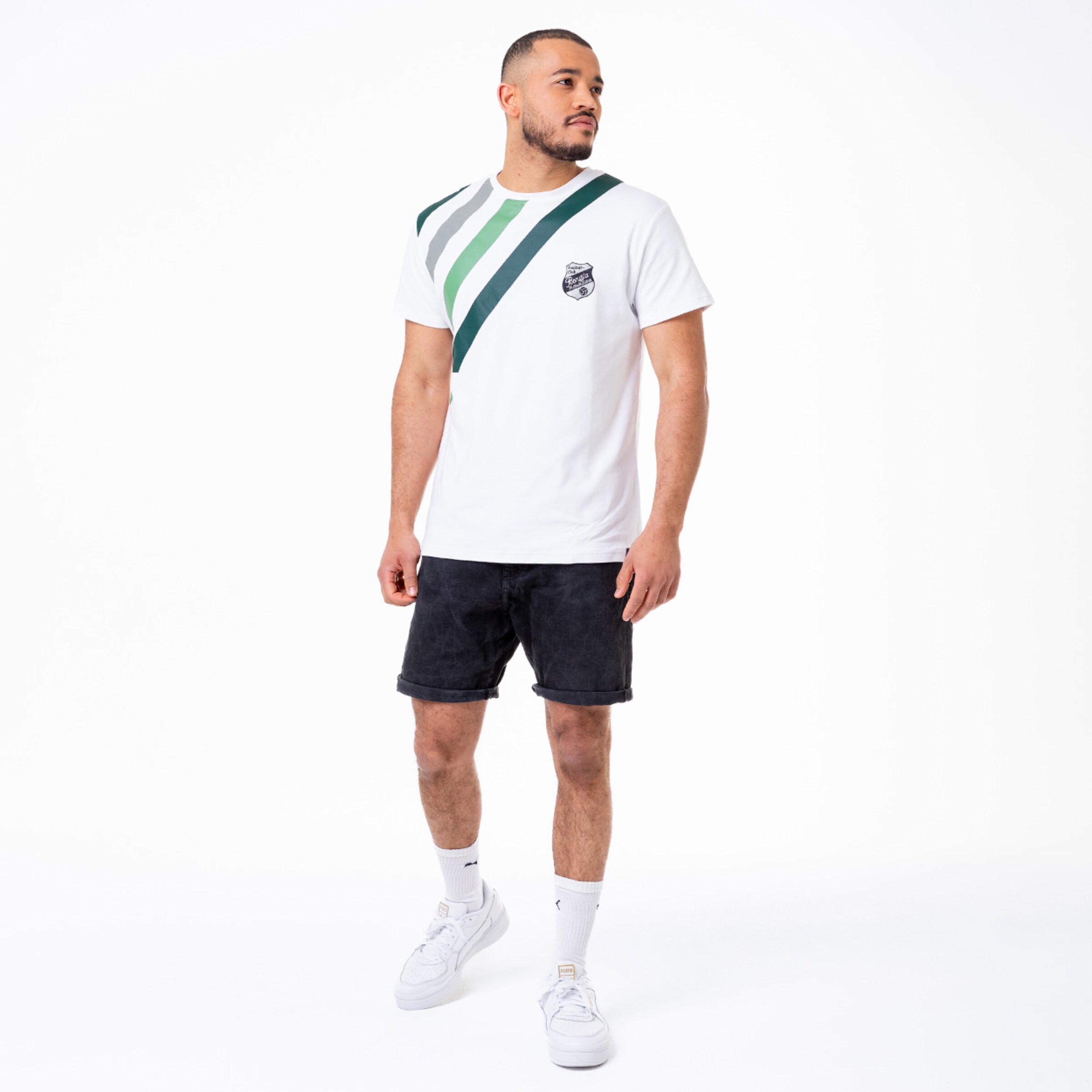 Mann in weißem T-Shirt mit diagonalen grünen Streifen und Logo kombiniert mit schwarzen Shorts und weißen Sneakers.