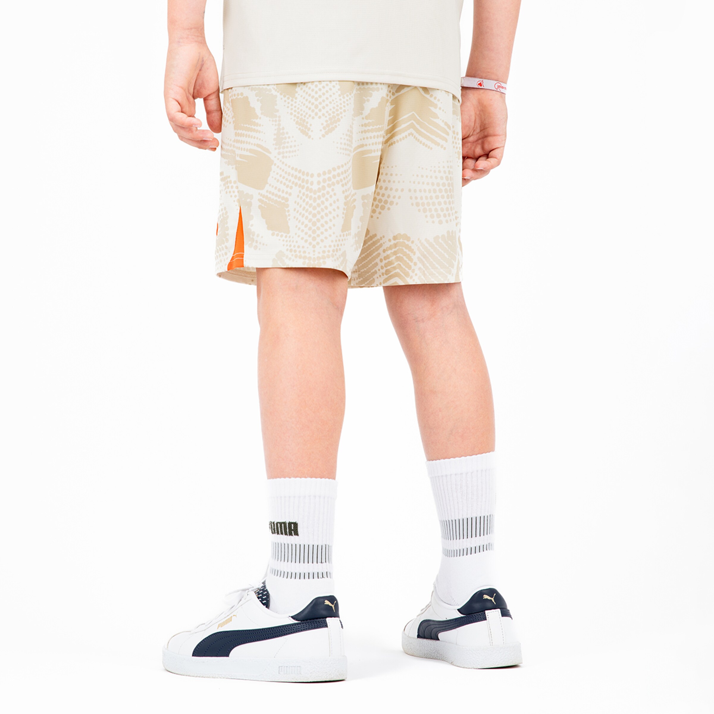 A boy is wearing beige sports shorts with a graphic pattern white socks and Puma sneakers.