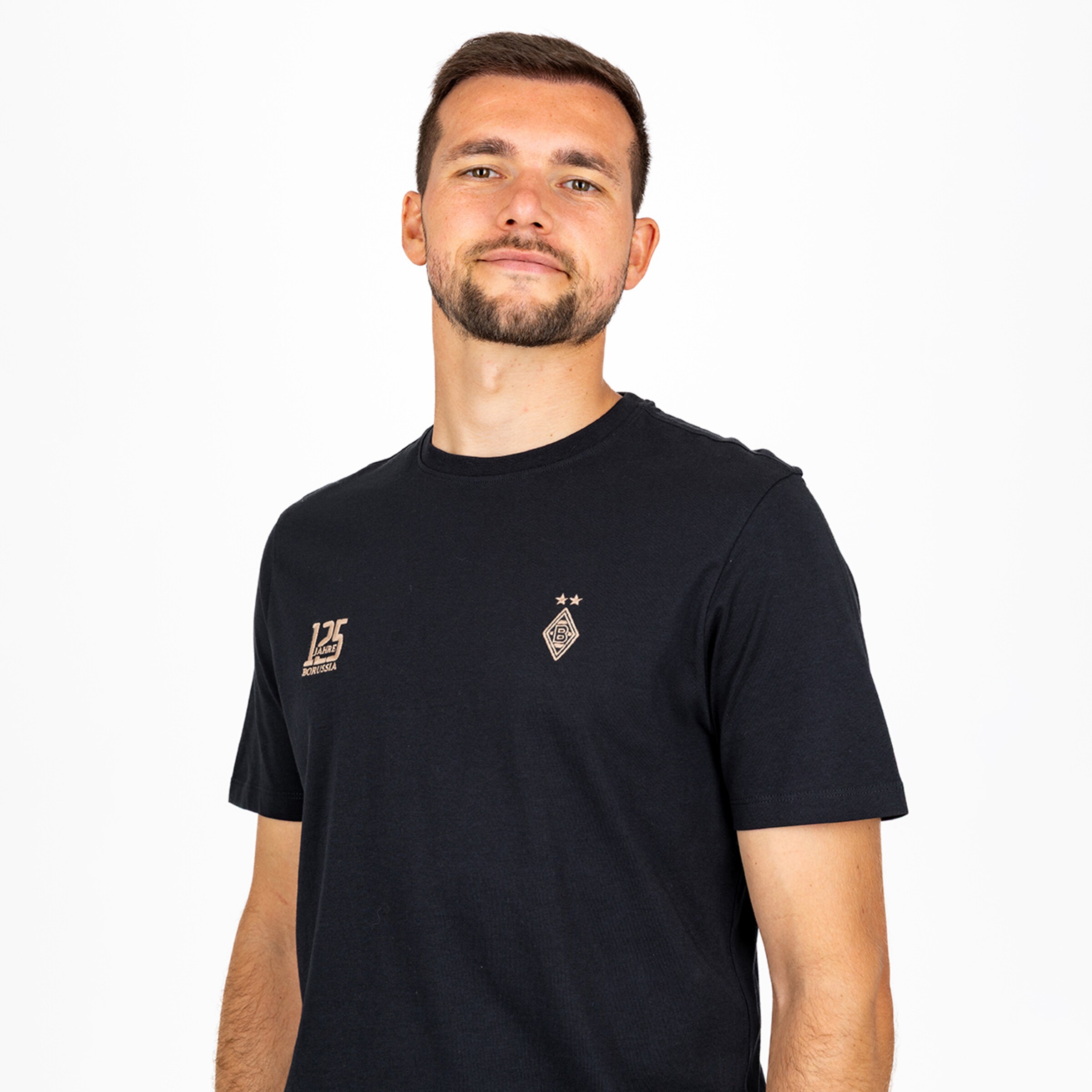 Ein Mann mit kurzen braunen Haaren und Bart trägt ein schwarzes T-Shirt mit zwei goldenen Logos vor einem weißen Hintergrund.