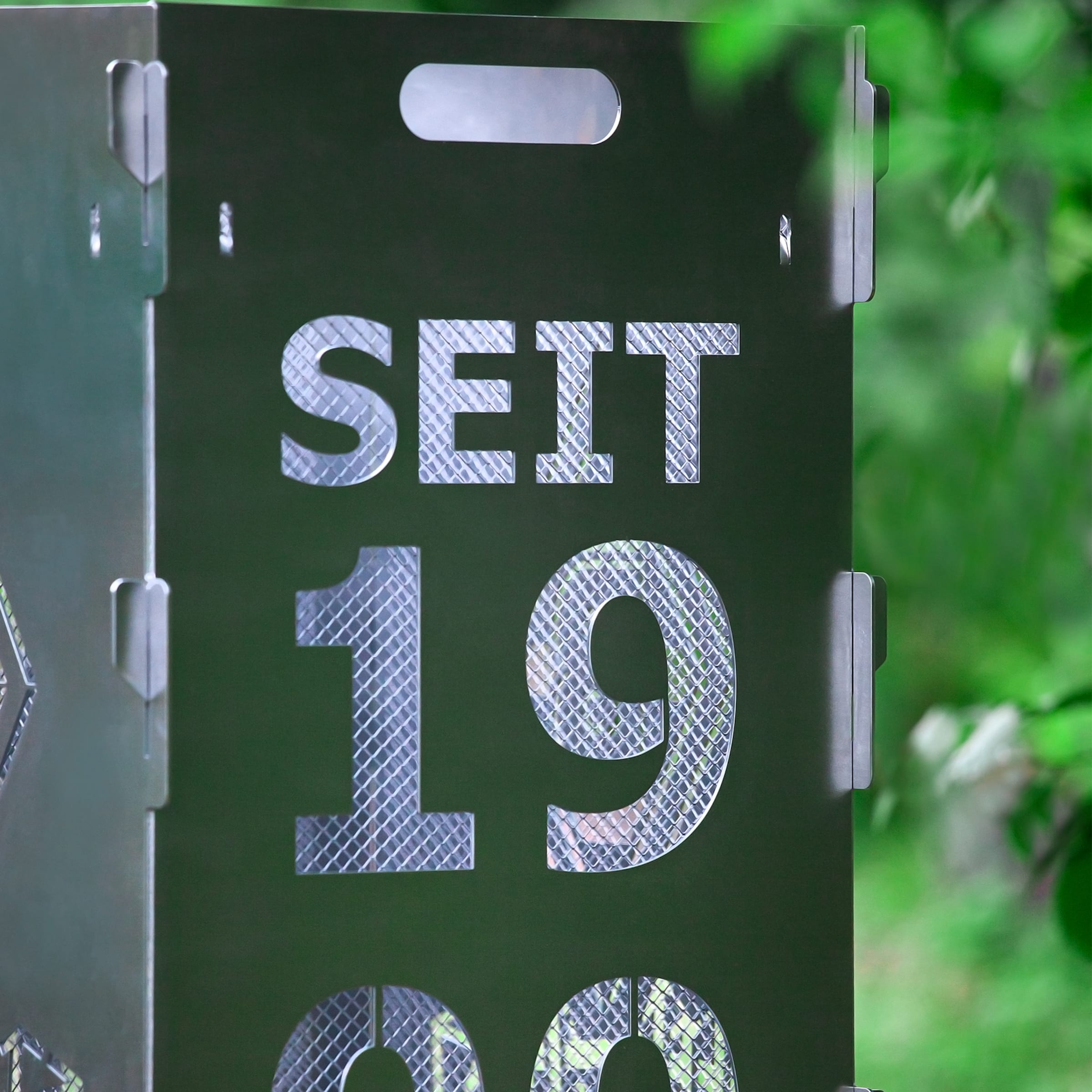 Nahaufnahme einer dunklen Metallstele mit dem ausgestanzten Text „SEIT 1990“ vor grünem Hintergrund. Ein Gitter schimmert durch die Buchstaben.