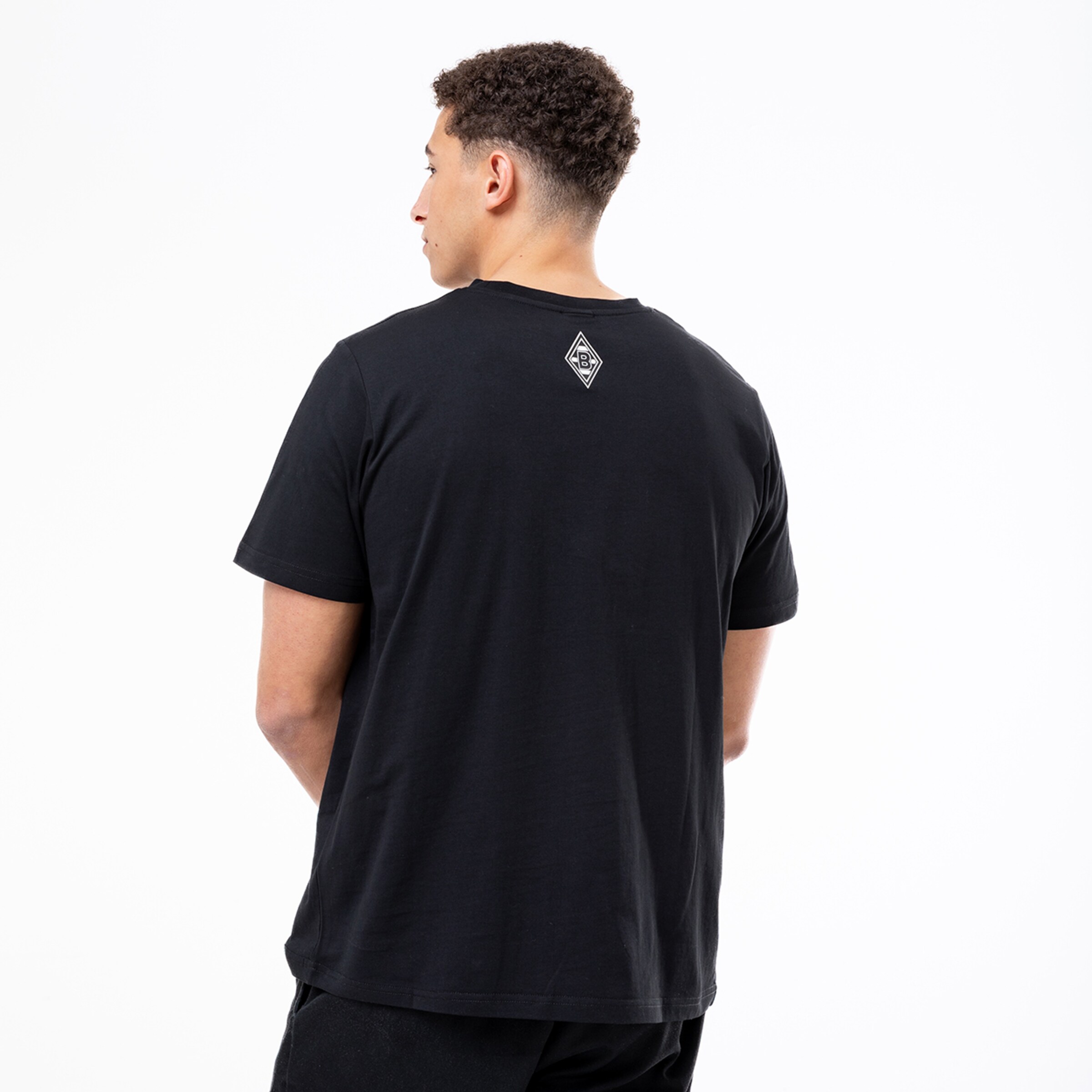 Rückansicht eines Mannes in einem schwarzen T-Shirt mit kleinem weißen Rauten-Logo im Nacken vor weißem Hintergrund.