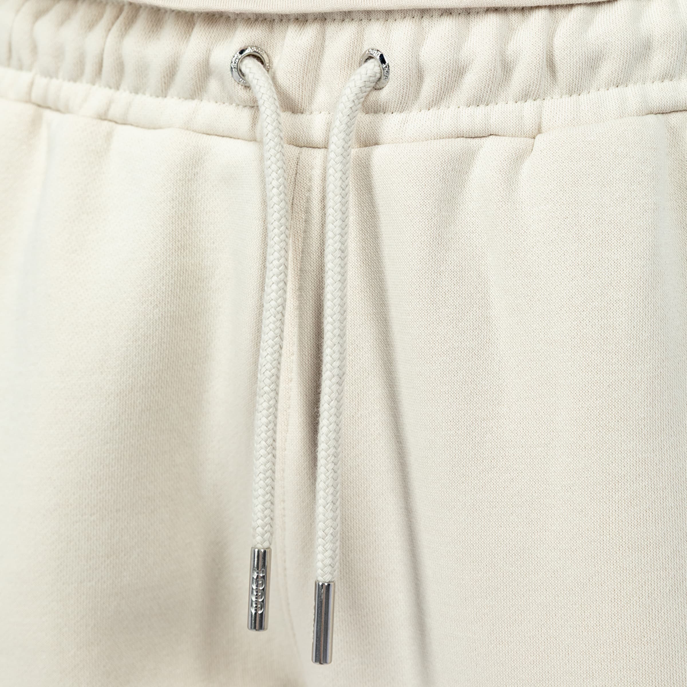 Close-up of cream-colored sweatpants showing an elastic waistband and braided drawstring with silver metal tips. The fabric has a soft, knitted texture.