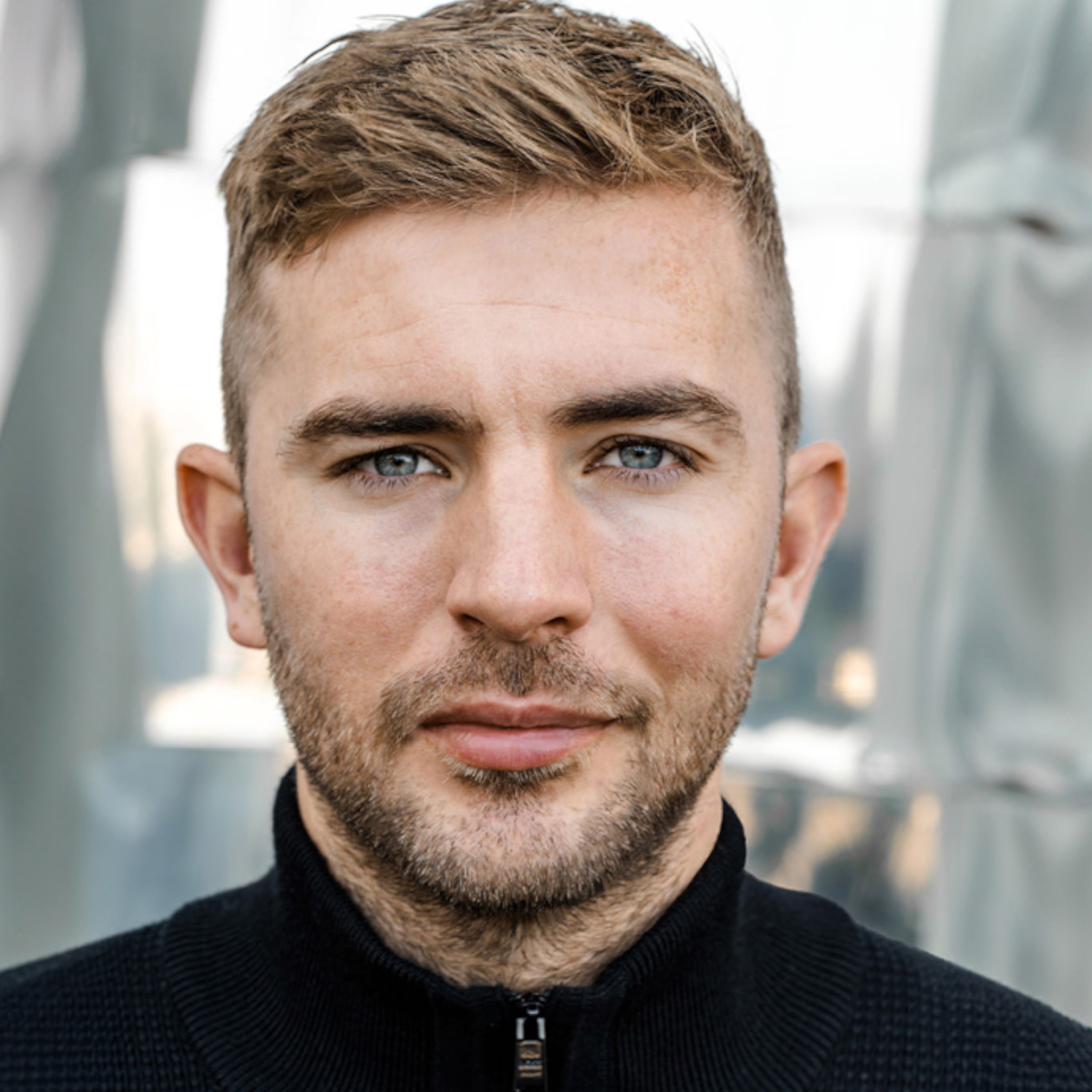 Porträt eines Mannes mit blondem Haar, blauen Augen und Bartstoppeln in einem schwarzen Reißverschlusspullover vor hellem Hintergrund.