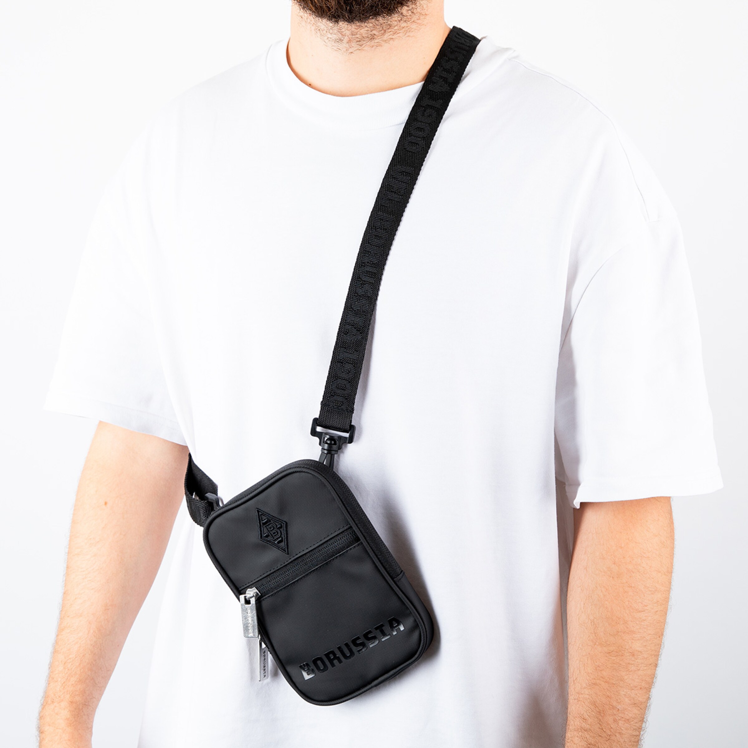 Schwarze Umhängetasche mit Schultergurt getragen von einer Person die ein weißes T-Shirt trägt.