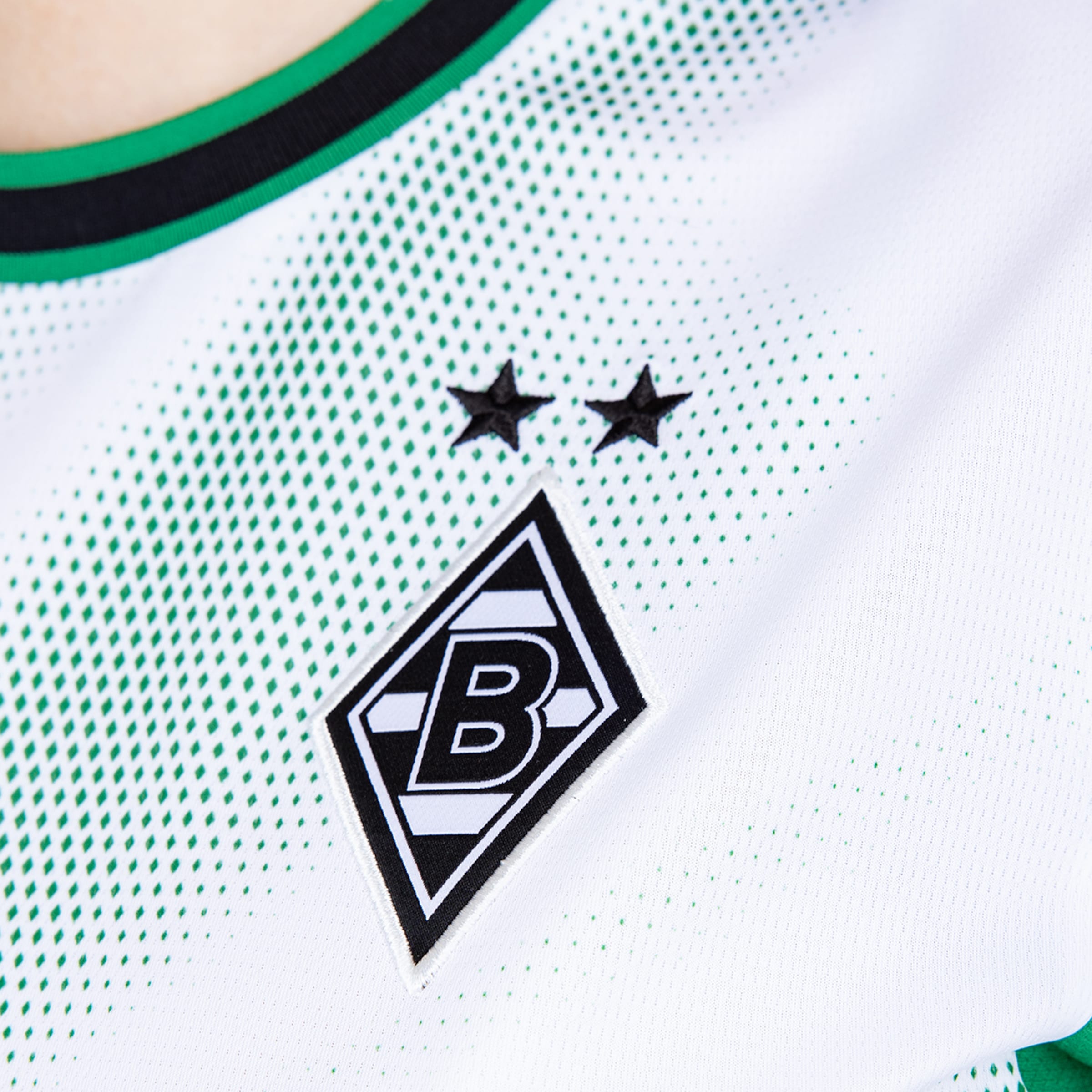 Nahaufnahme eines weißen Borussia Mönchengladbach Trikots mit dem Vereinslogo und zwei Sternen auf einem Muster aus grünen Punkten.