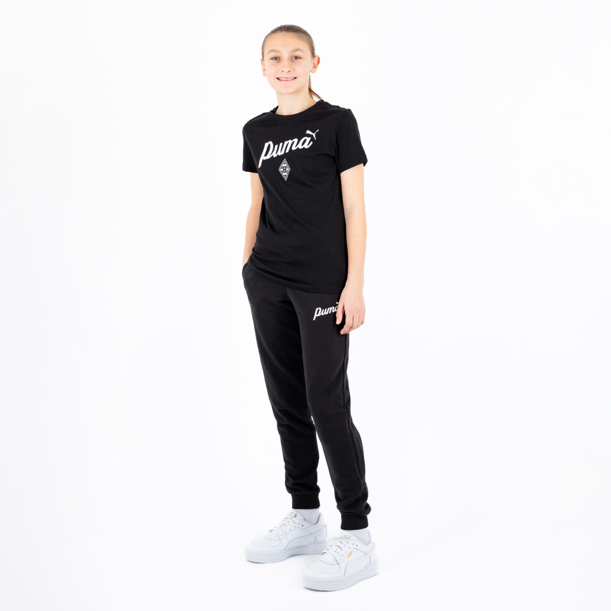 A girl stands in athletic clothing wearing a black t-shirt and sweatpants with white shoes.