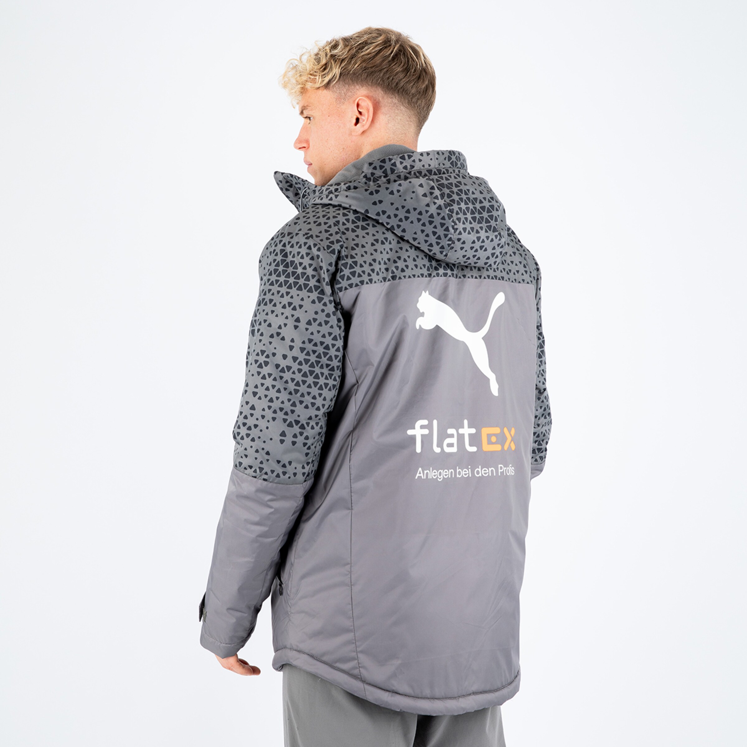 Junger Mann mit blondem Haar trägt eine graue Jacke von Puma mit Muster auf den Ärmeln und Logo auf dem Rücken.
