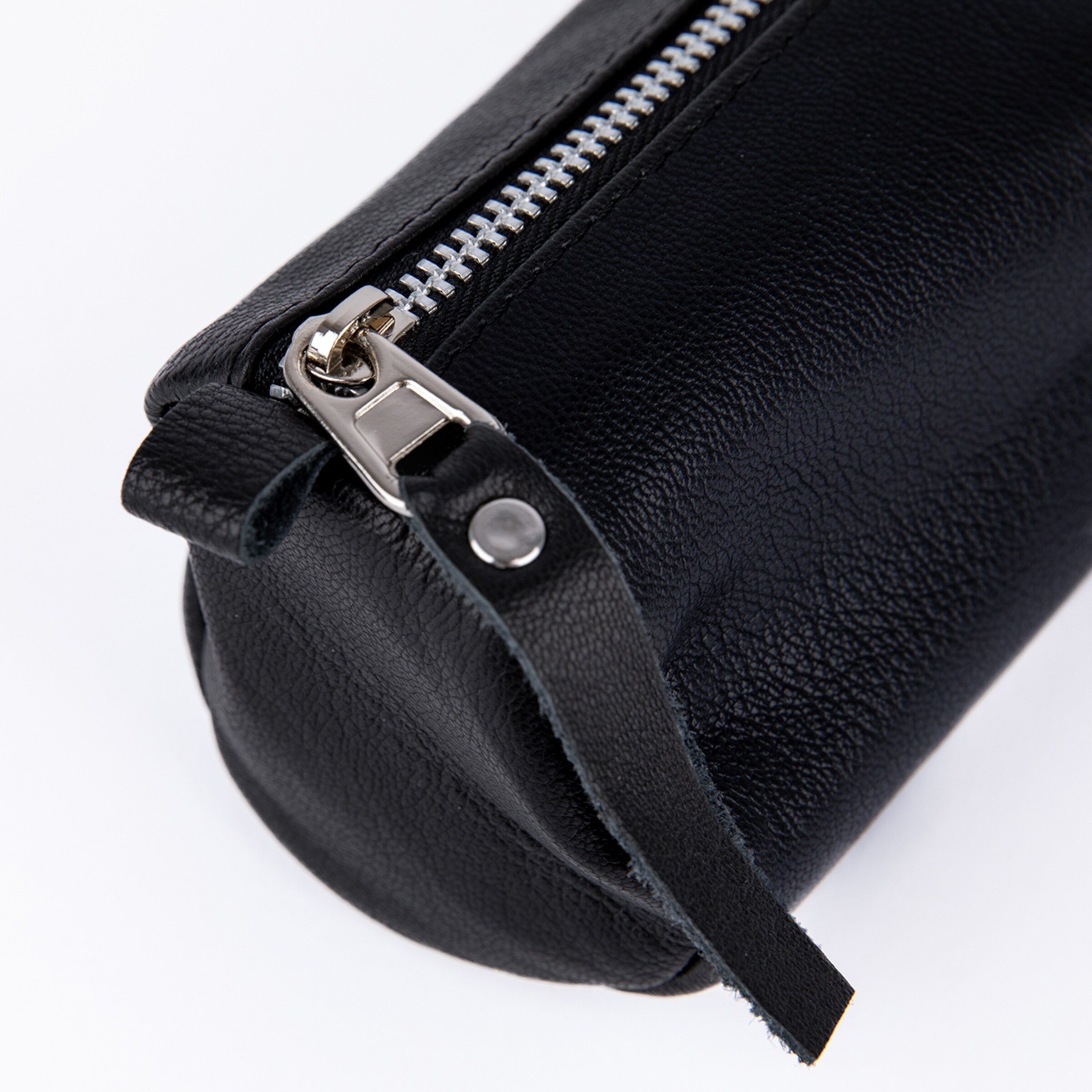Close-up of a black textured leather cylindrical pouch with a silver zipper and a leather pull tab secured by a silver rivet, set against a white background.