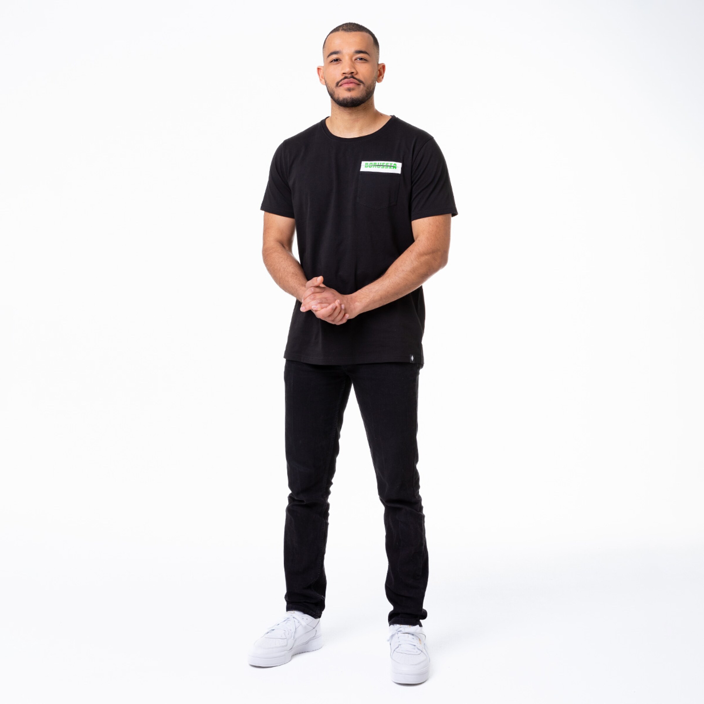 Mann in schwarzem T-Shirt und Jeans mit weißen Sneakern steht mit verschränkten Händen.