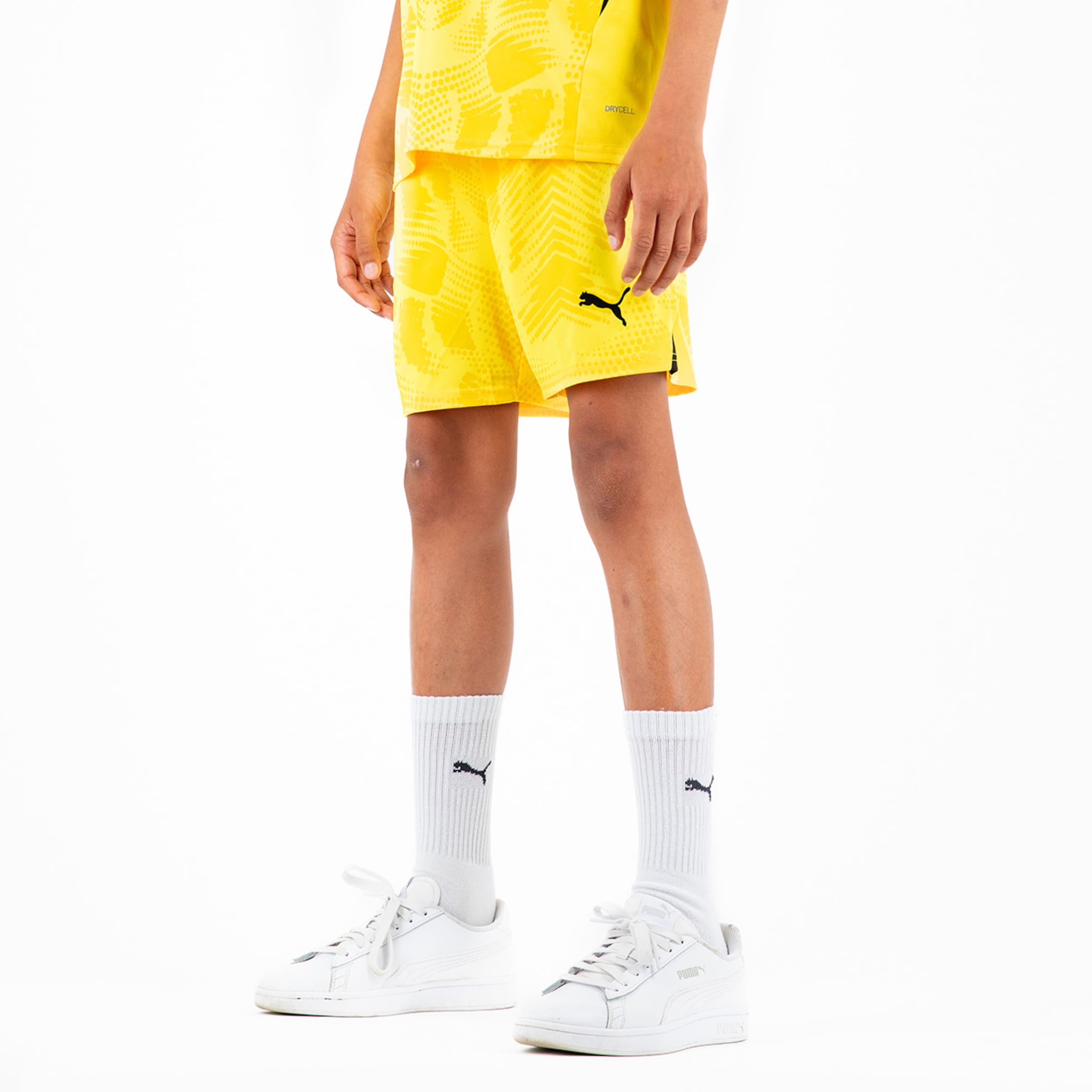 Junge trägt gelbes Sportoutfit mit kurzen Hosen und weißen Sneakers.