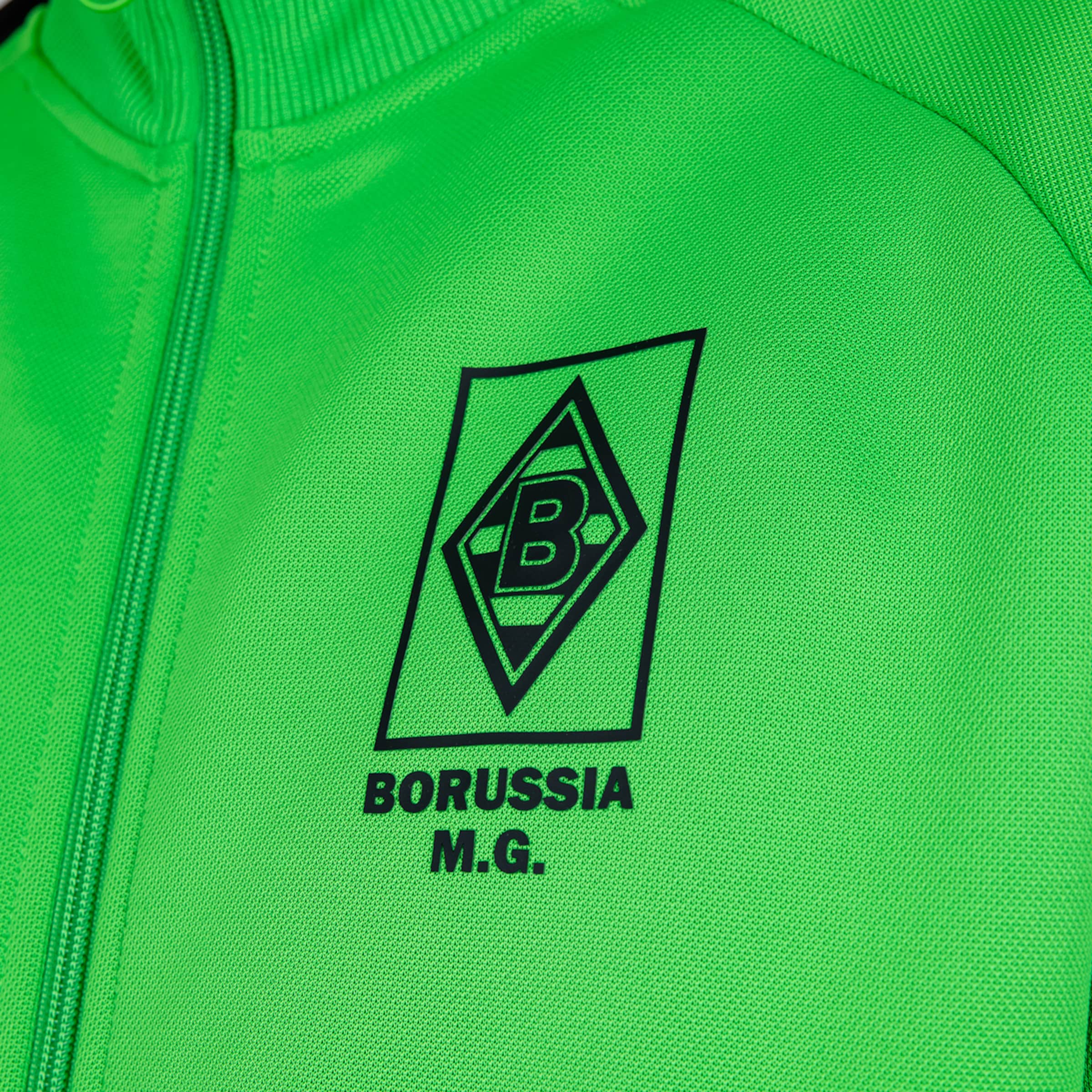 Nahaufnahme einer neongrünen Trainingsjacke mit dem schwarzen Logo von Borussia Mönchengladbach und dem Schriftzug "BORUSSIA M.G.".
