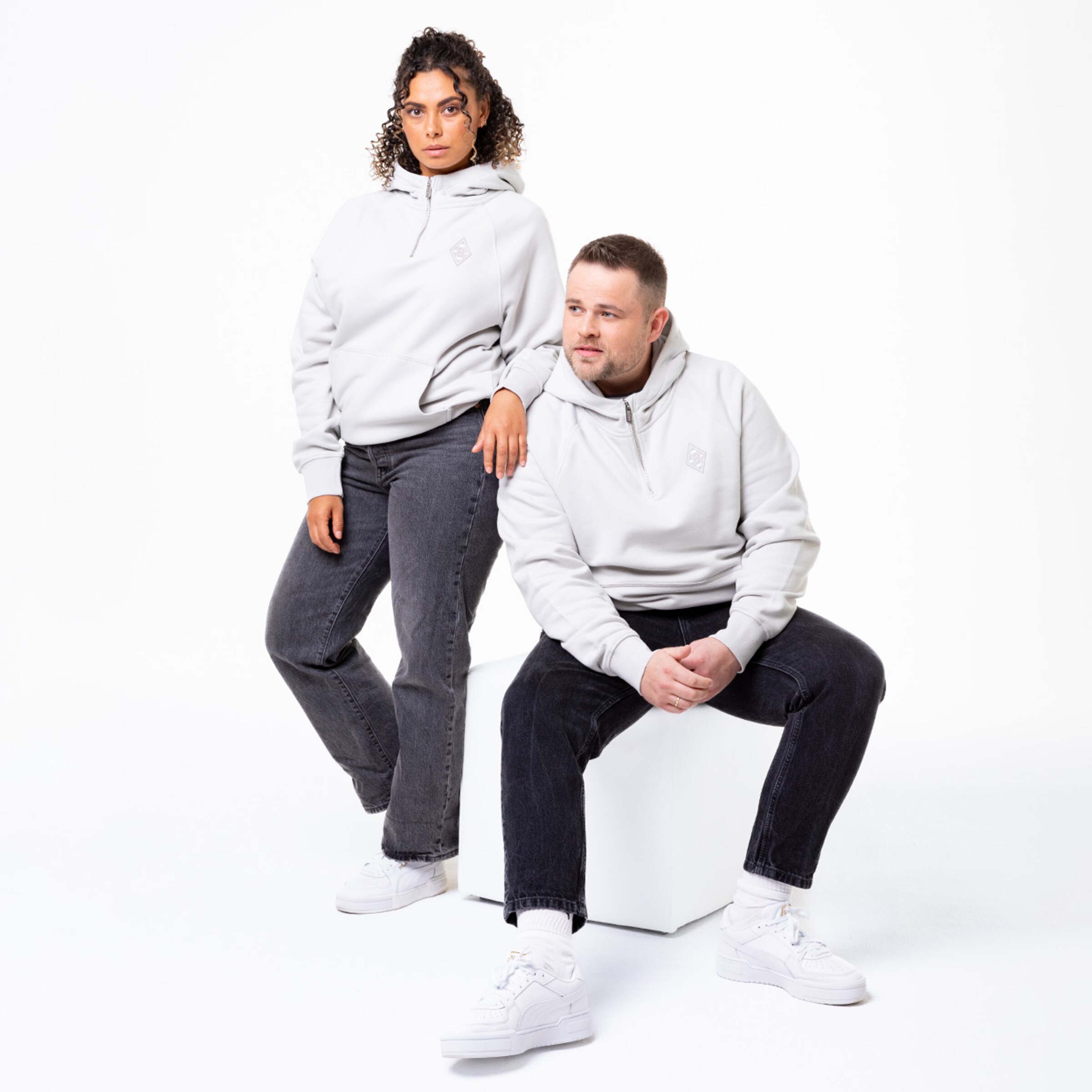 Zwei Personen in grauen Hoodies und schwarzen Jeans stehen und sitzen auf einem weißen Würfel.