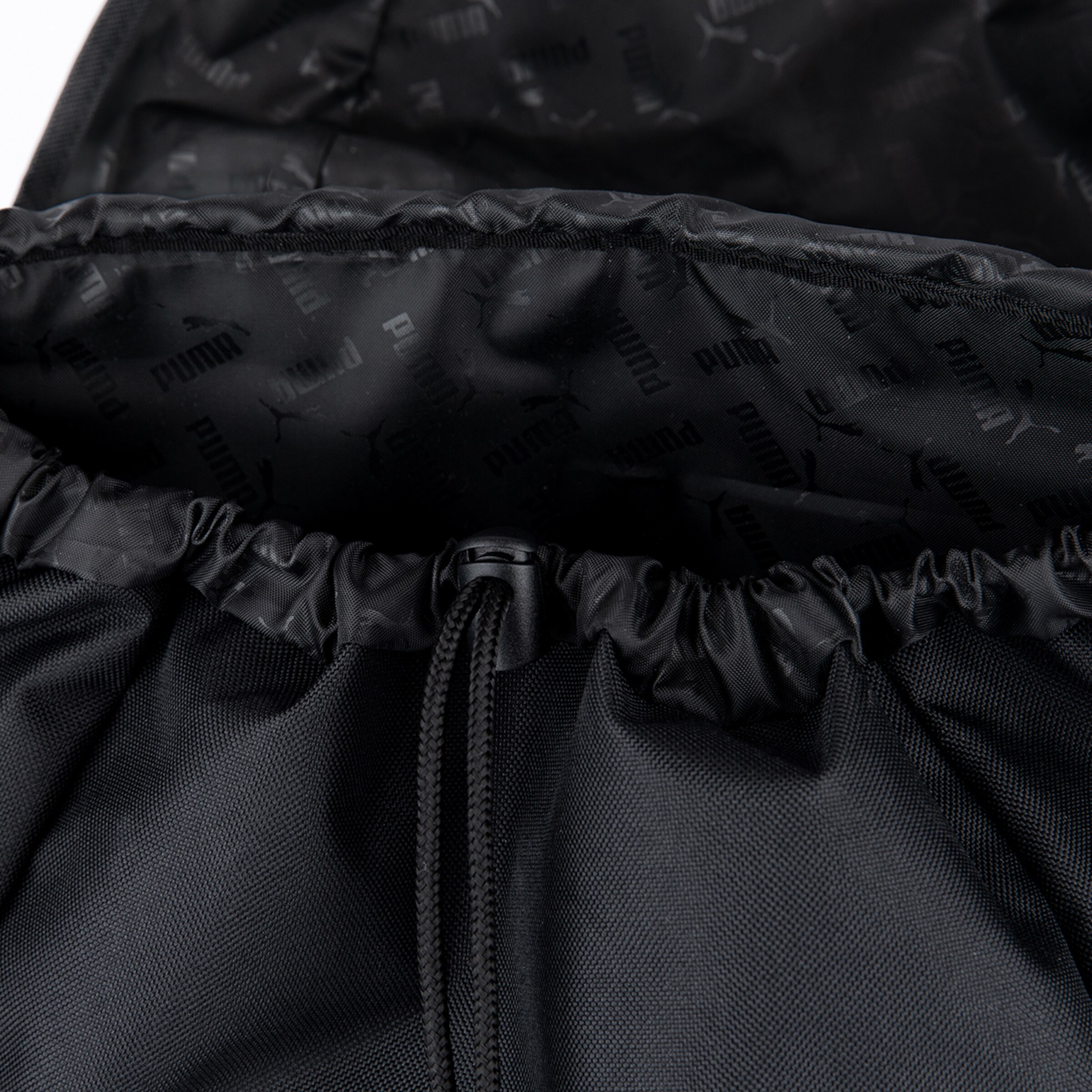 Nahaufnahme des Innenraums einer schwarzen Beuteltasche, die ein Futter mit dezenten, wiederholten Puma-Logos und -Schriftzügen zeigt. Der schwarze Webstoff und der Kordelzug mit Stopper sind oben sichtbar.
