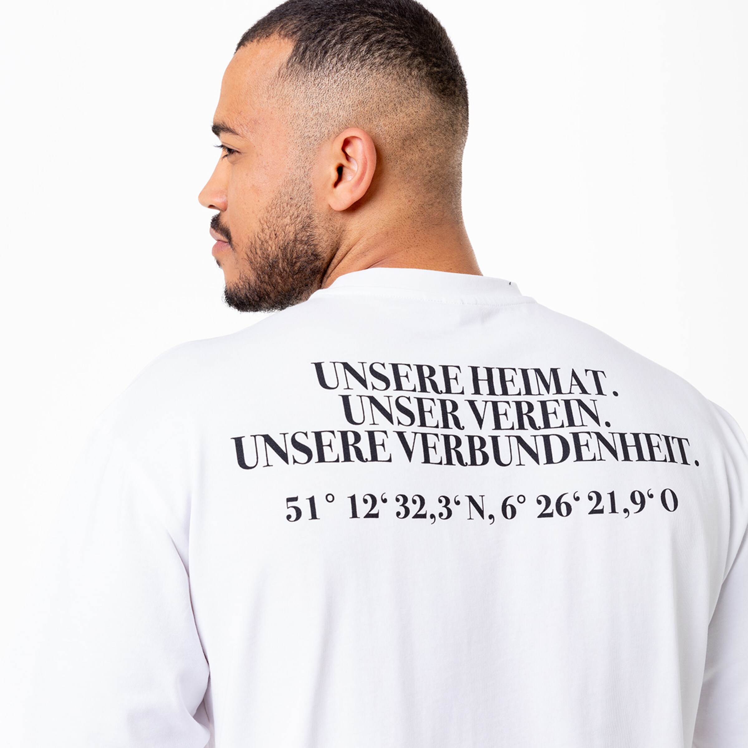 Mann mit Rucksack trägt ein weißes Shirt mit Aufschrift auf dem Rücken: UNSERE HEIMAT. UNSER VEREIN. und Koordinaten.