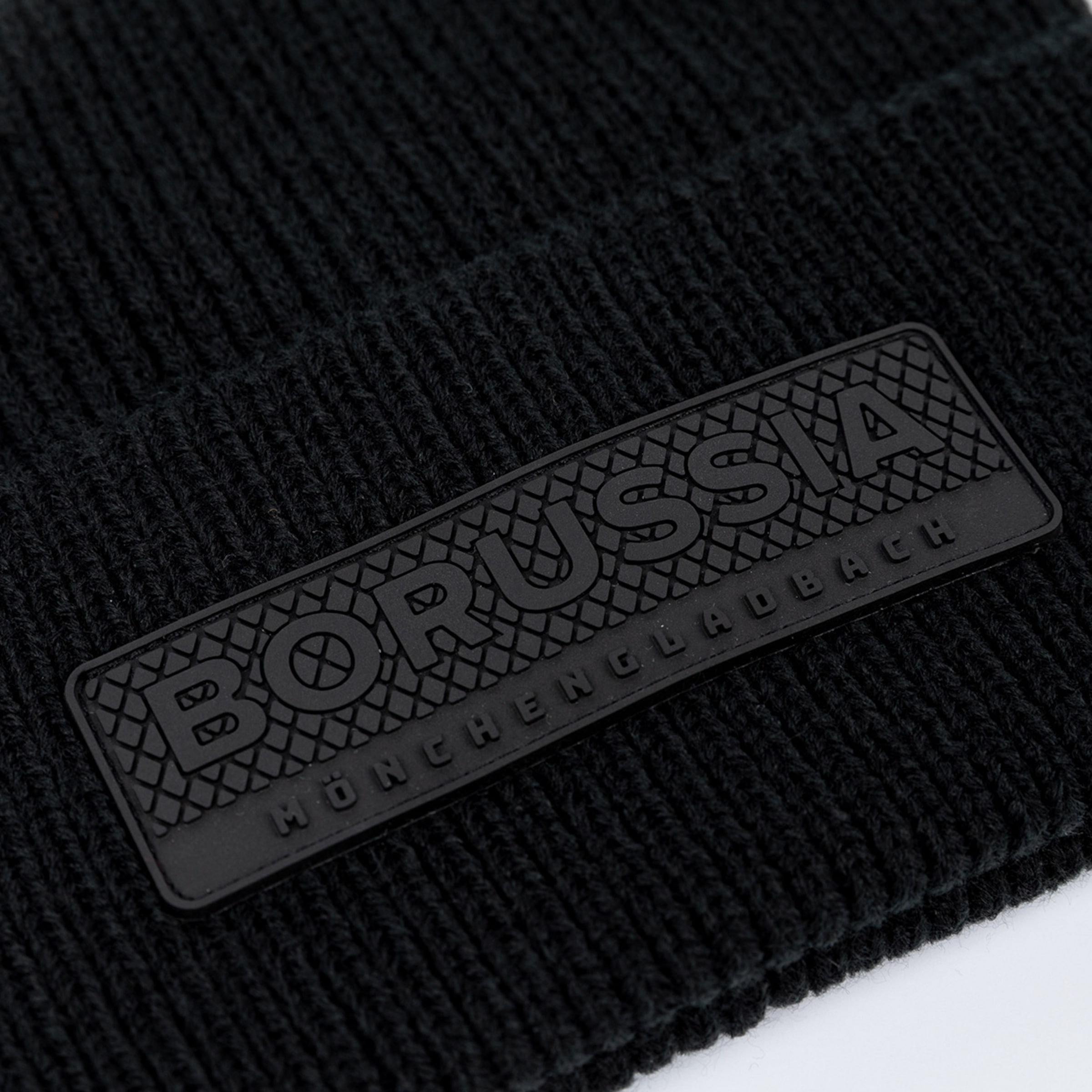 Close-up of a black knitted beanie with an embossed rubber patch featuring "BORUSSIA MÖNCHENGLADBACH" on a textured background.