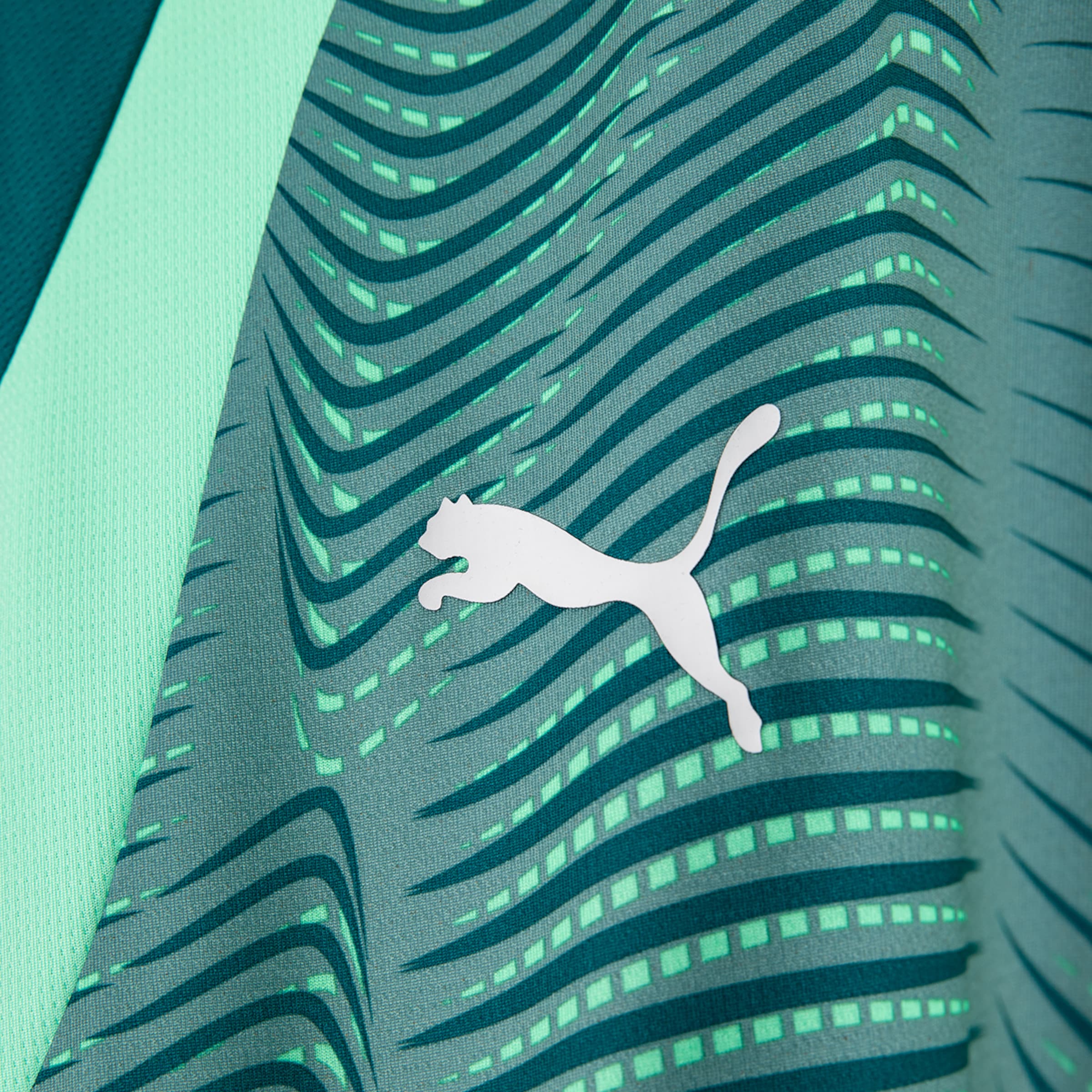 Close-up of a PUMA sports jersey featuring the leaping Puma logo in white against a background of wavy green stripes and rectangular light green segments on teal fabric. A strip of light green fabric is visible on the left.