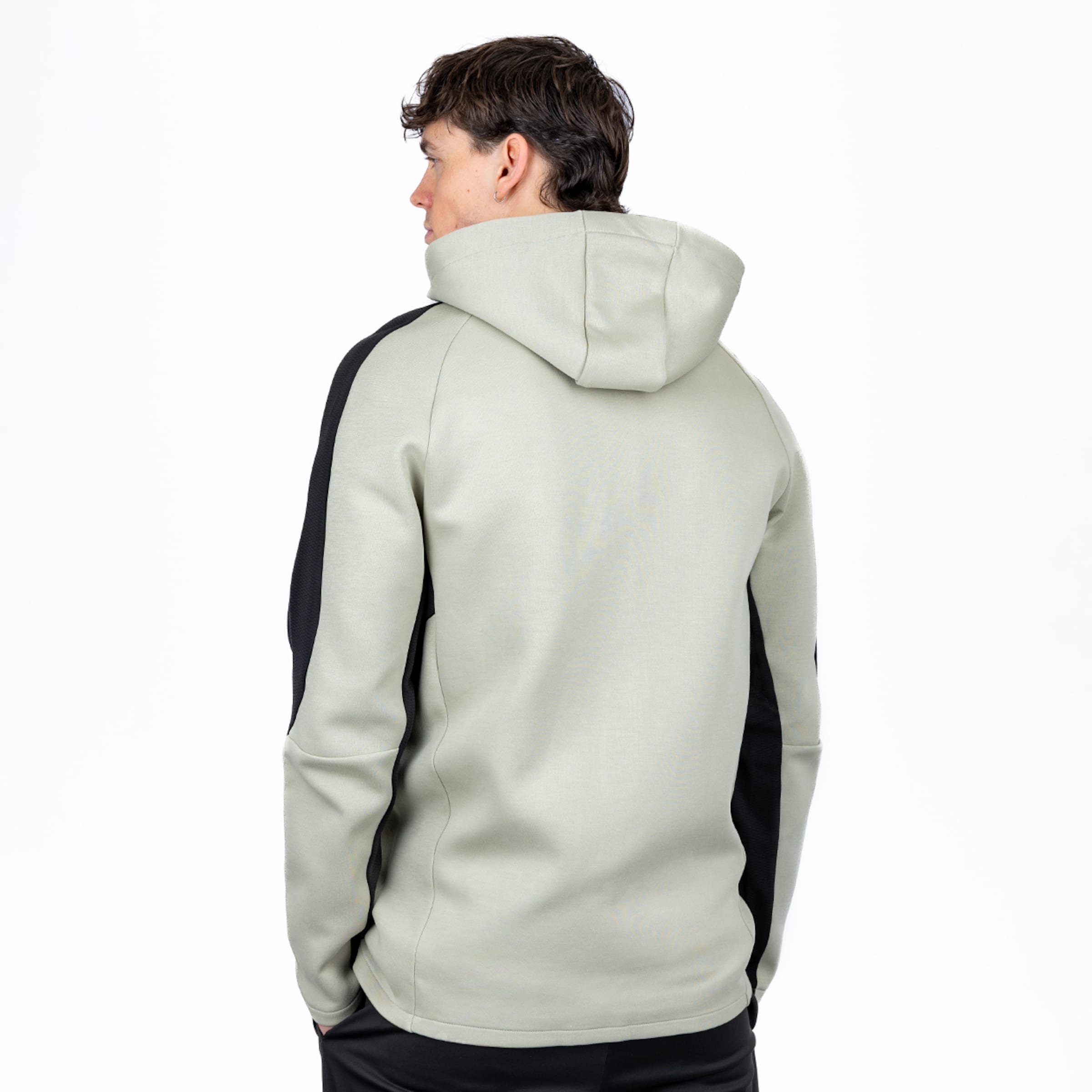 Rückenansicht eines Mannes in einem hellen salbeigrünen Hoodie mit schwarzen Seiteneinsätzen, der über seine linke Schulter blickt. Er hat dunkles, welliges Haar und einen silbernen Ohrring.