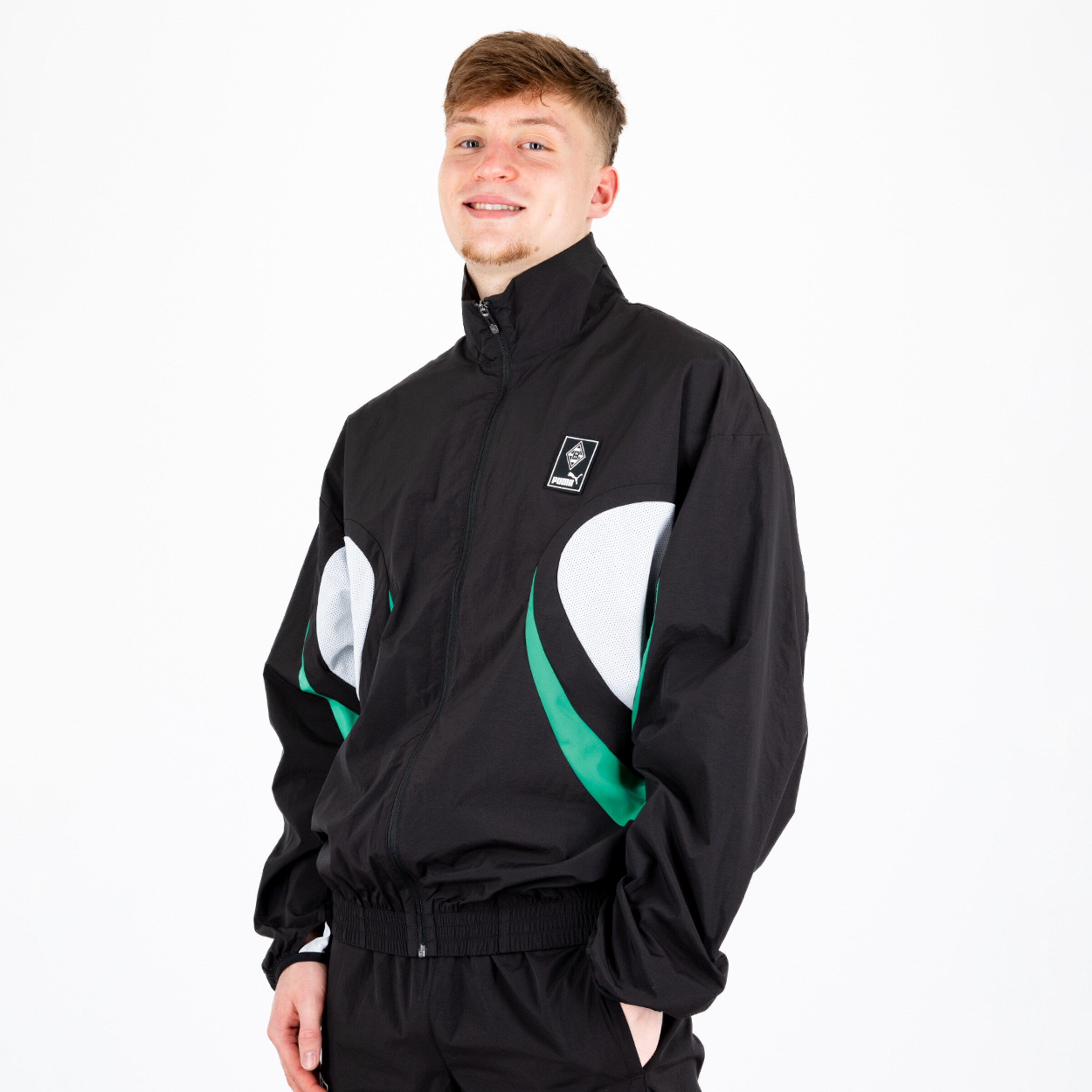 Junger Mann in schwarzer Jacke mit grünem Design lächelt und steht aufrecht.