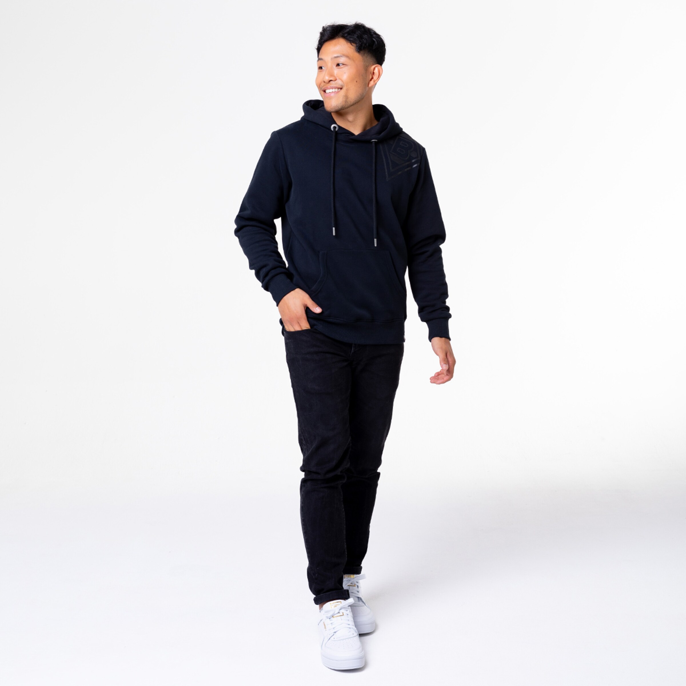 Mann trägt einen schwarzen Hoodie und schwarze Jeans geht lächelnd in eine Richtung.