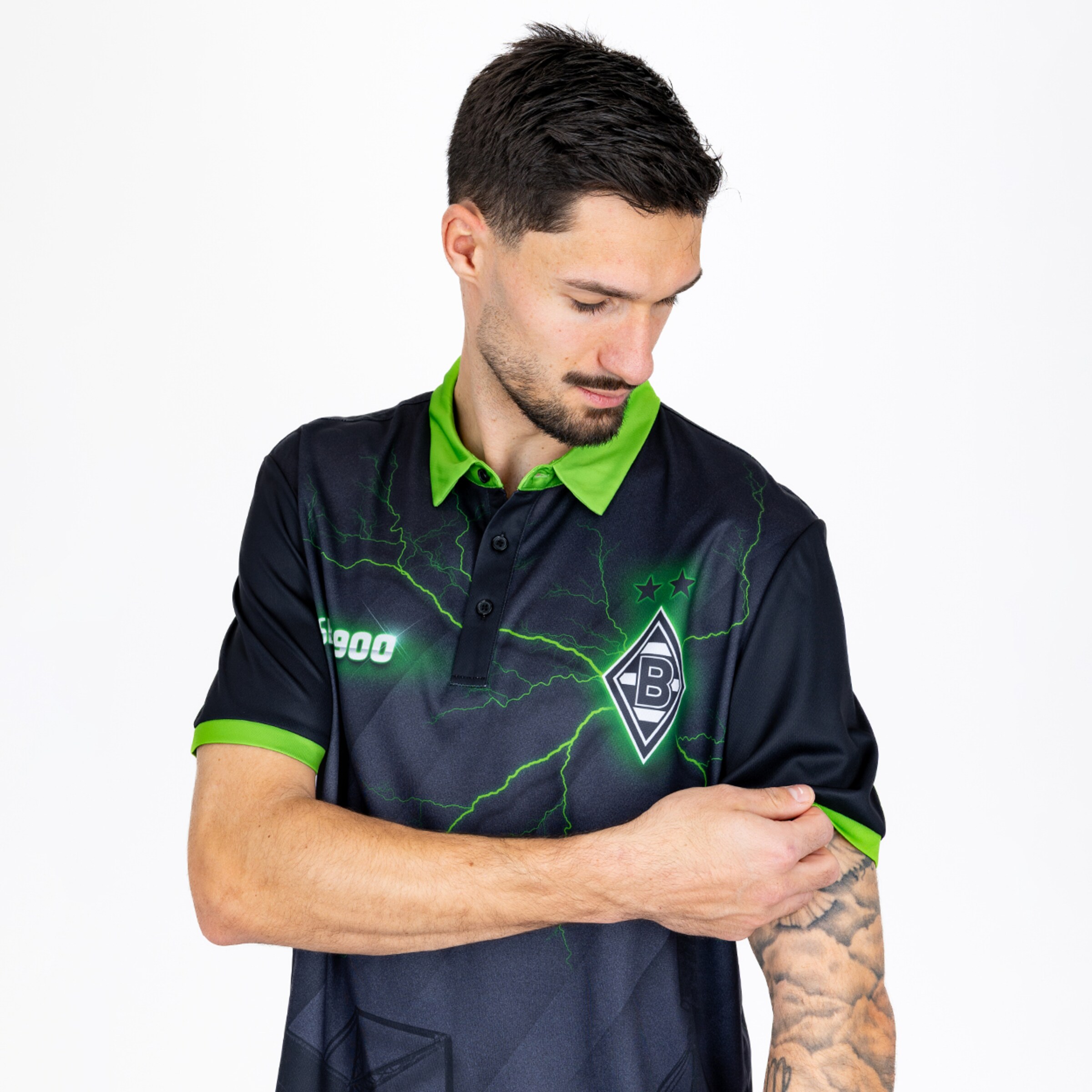 Mann im schwarzen Borussia Mönchengladbach Poloshirt mit grünen Blitzen, der seinen Ärmel hochkrempelt und ein Tattoo zeigt.