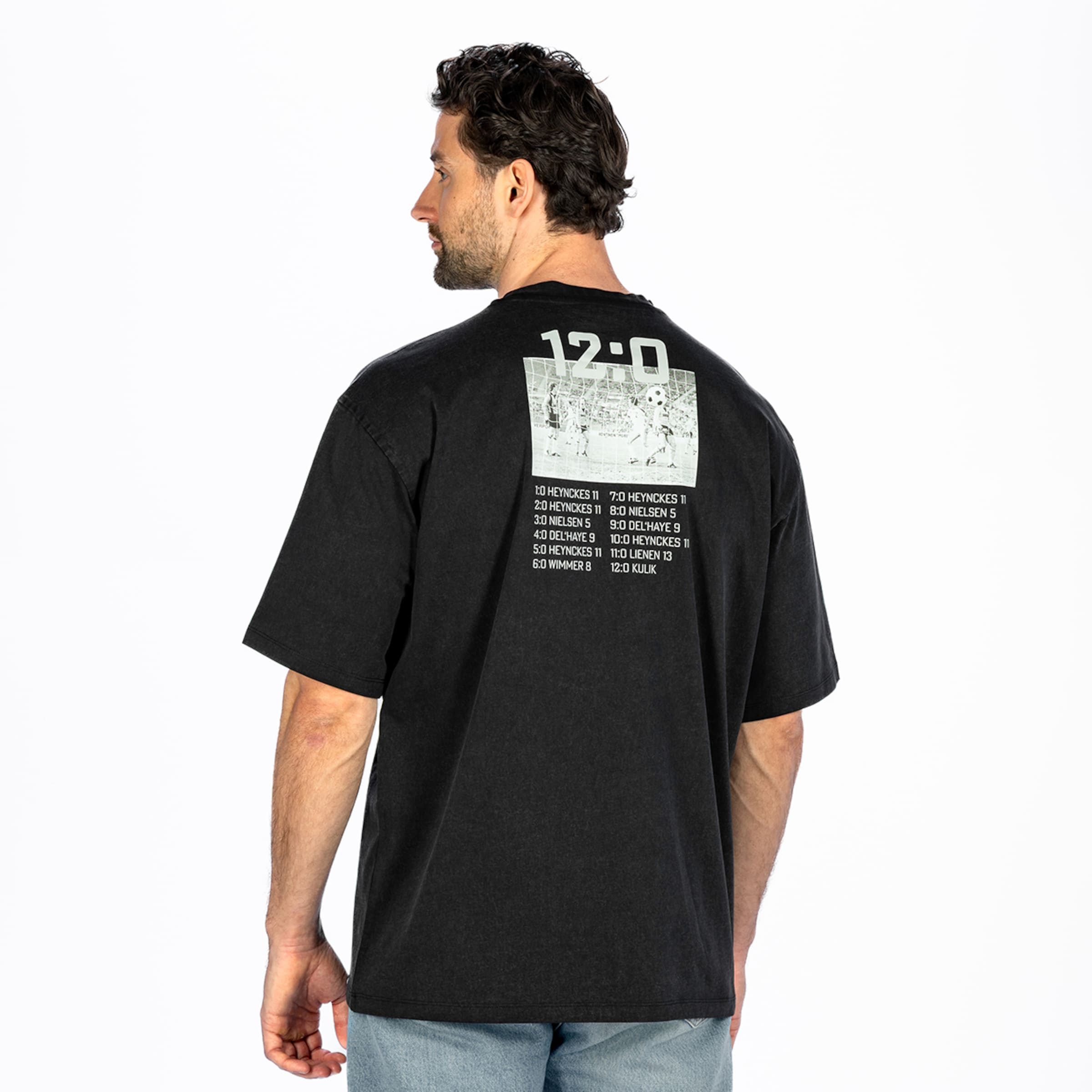 Back view: Man wearing a black oversized t-shirt with a "12:0" back print, goalscorer list and vintage football photo on a white background.