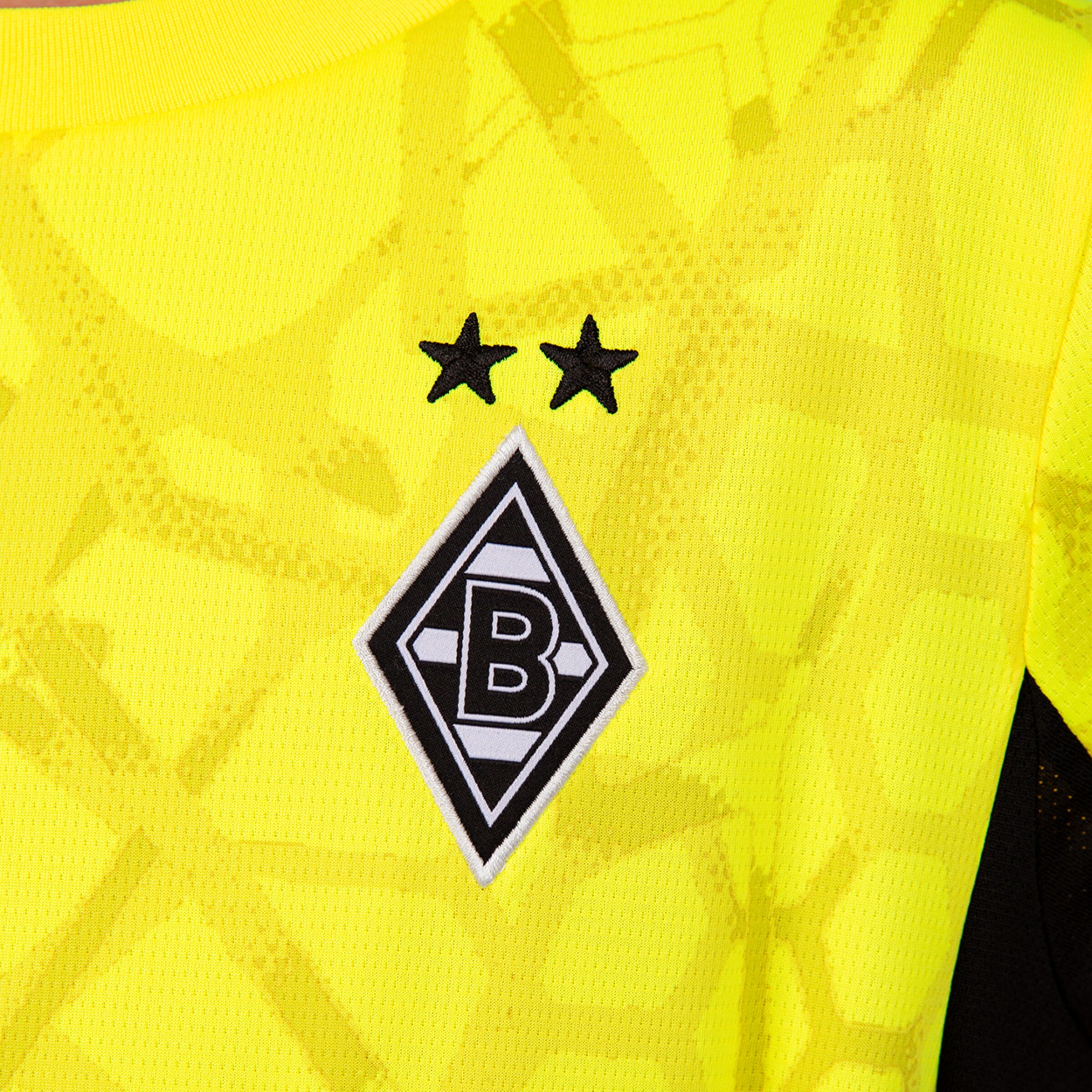 Nahaufnahme des Borussia Mönchengladbach Wappens mit zwei schwarzen Sternen auf einem neongelben Trikot mit Rautenmuster.