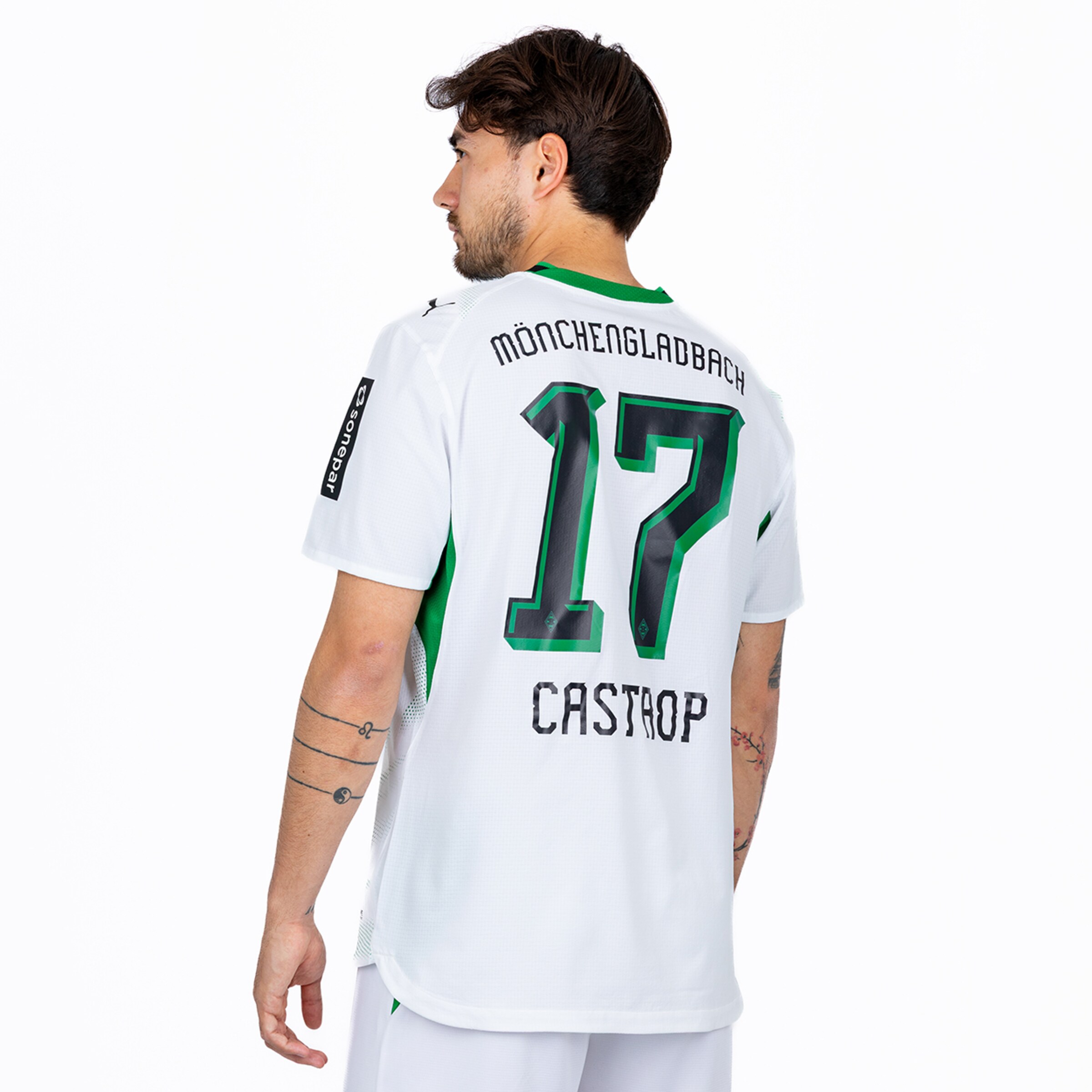 A man from behind, slightly turned, wears a white and green soccer jersey with 'MÖNCHENGLADBACH', '17', and 'CASTROP' in black/green on the back. Tattoos are visible on his arms.
