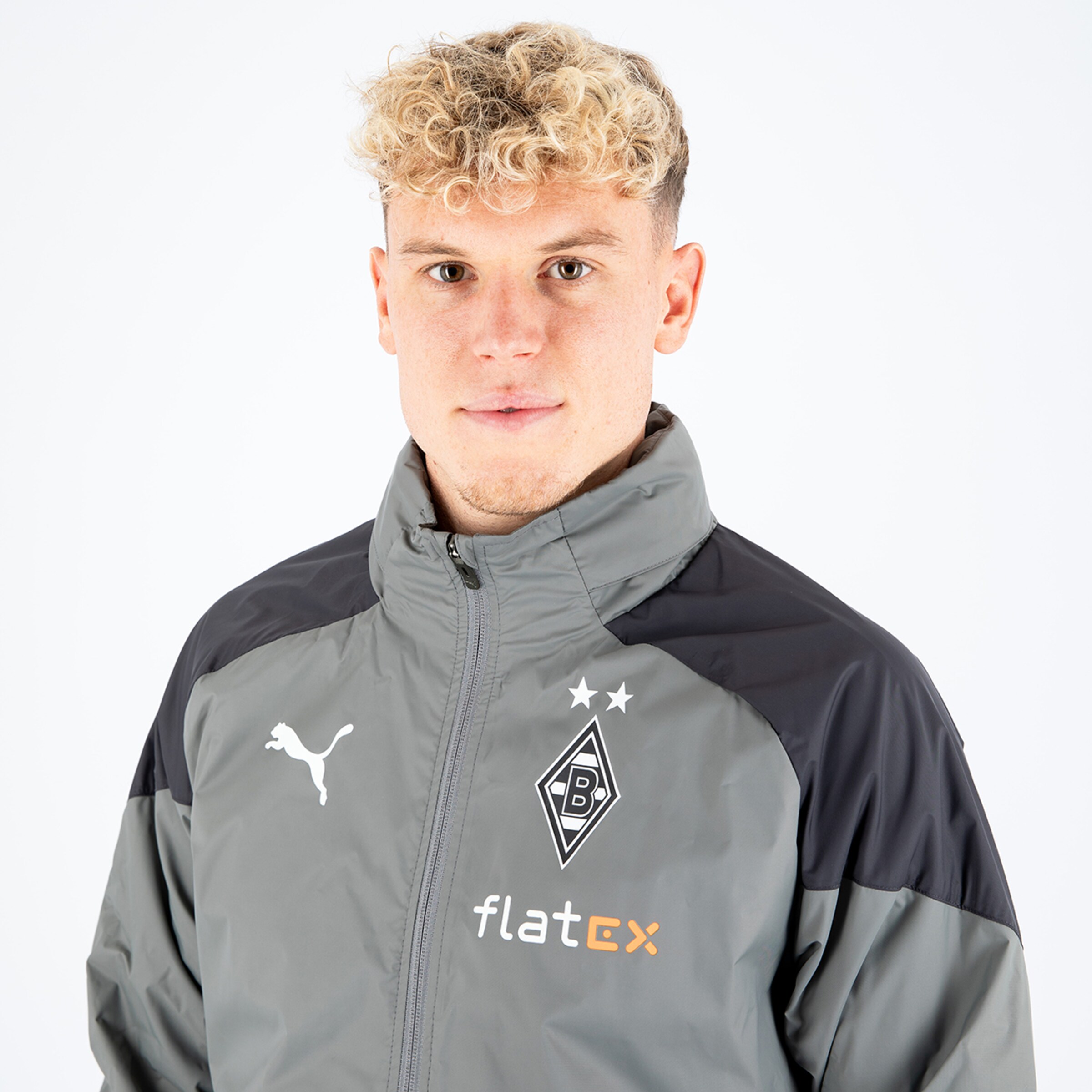 Junger Mann mit lockigem Haar trägt graue Jacke mit Puma-Logo und Borussia Mönchengladbach-Emblem.