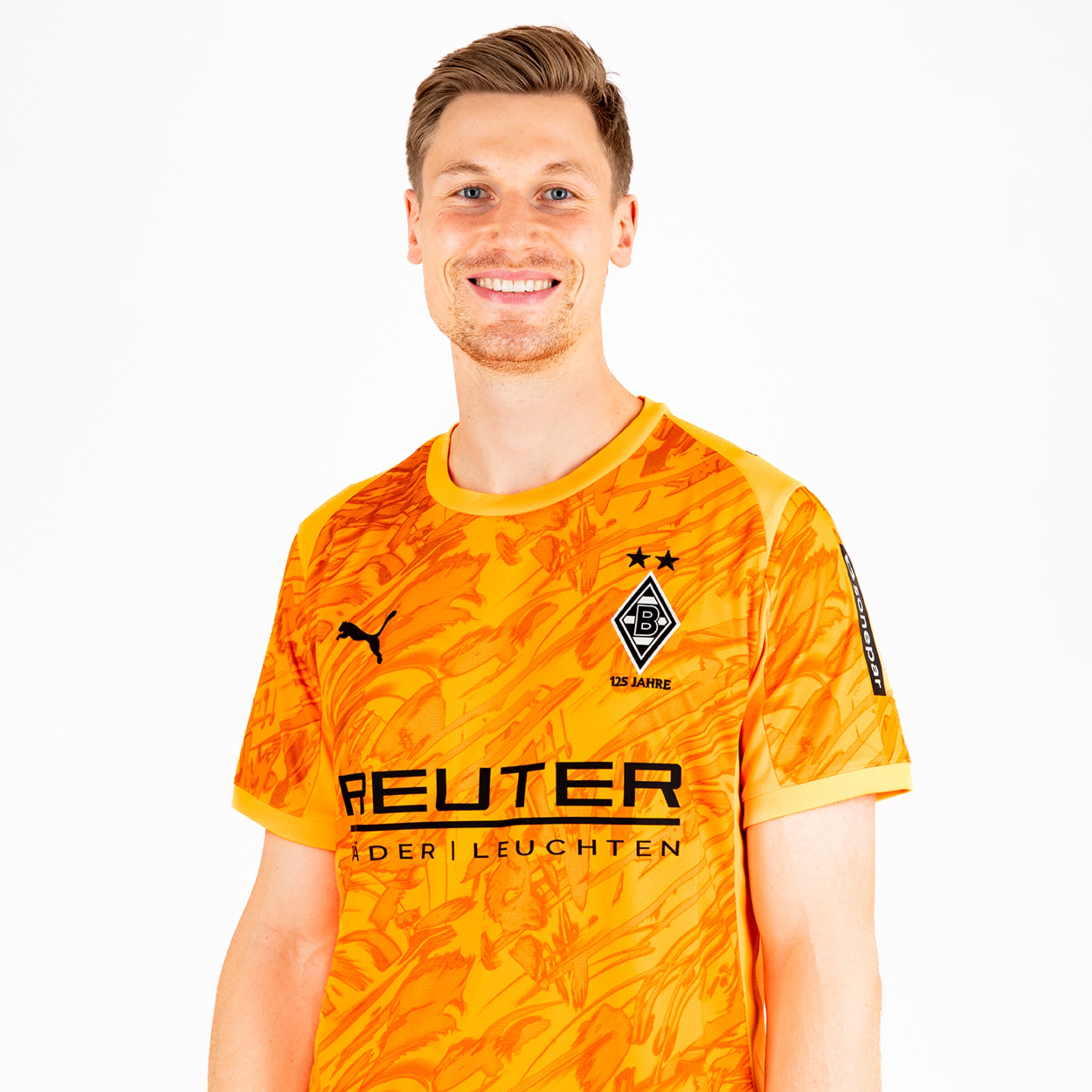 Ein Fußballspieler in einem orangefarbenen Trikot mit schwarzem Logo und Schriftzug lächelt freundlich.