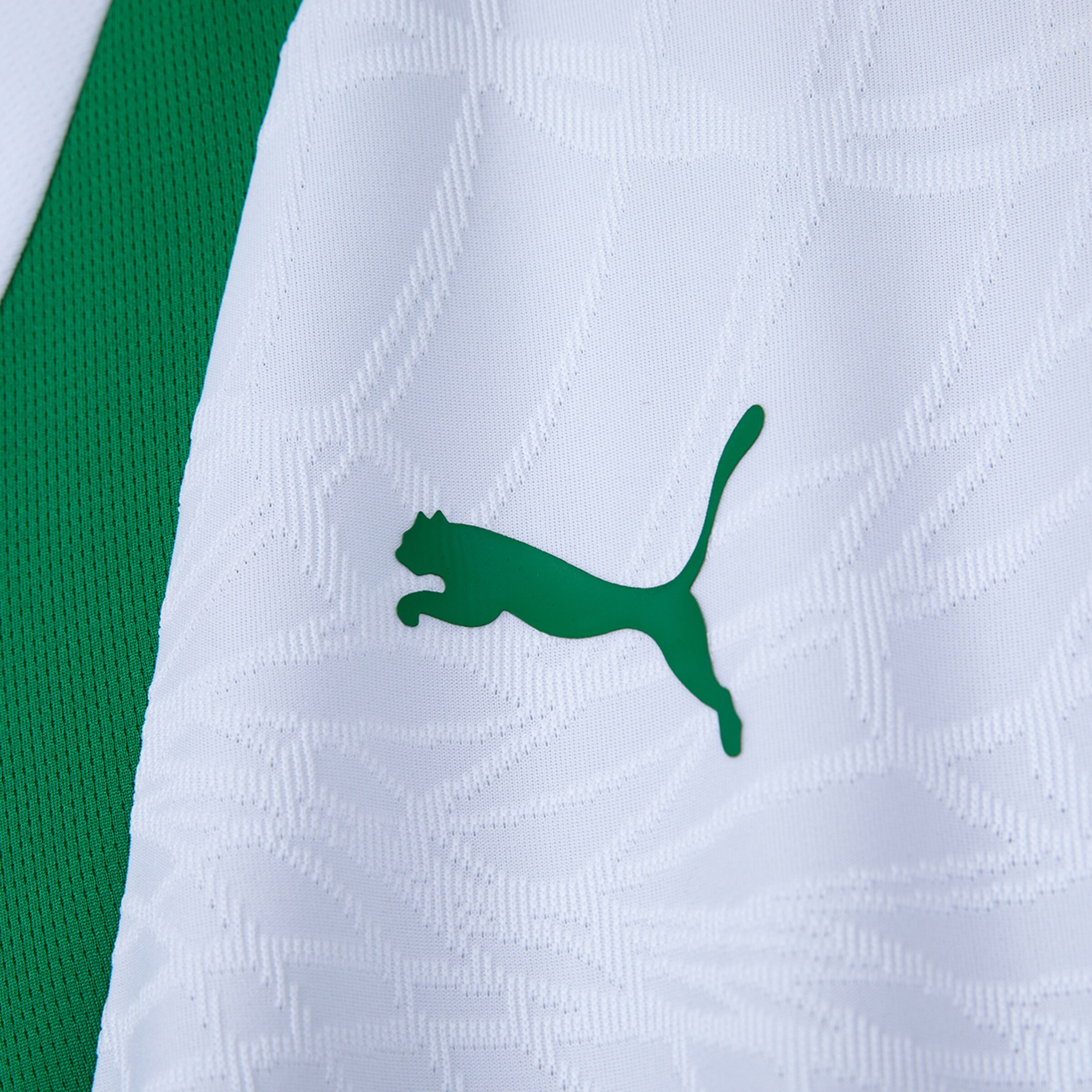 Close-up of a white sports jersey with a green Puma logo. The white fabric features an embossed pattern, next to a green mesh panel.