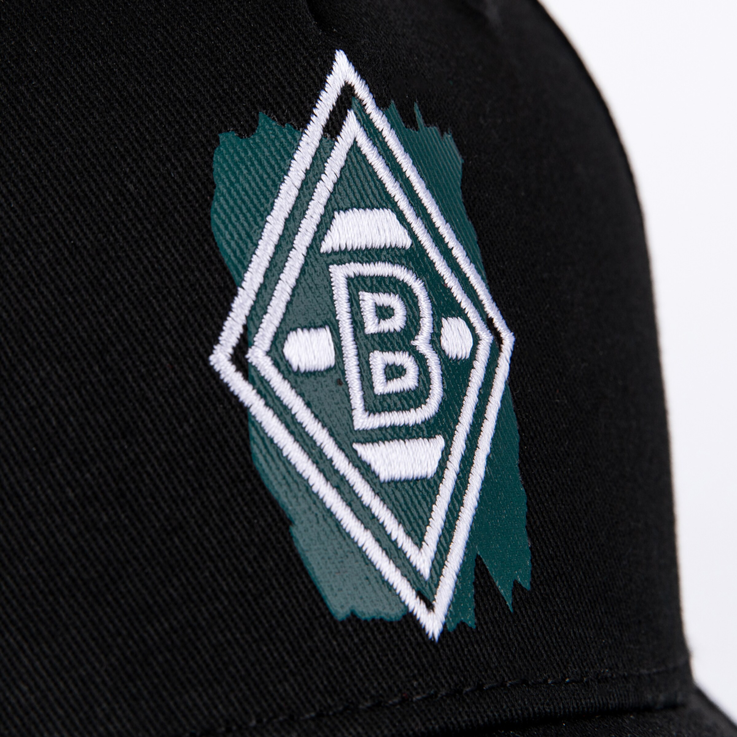Nahaufnahme einer schwarzen Kappe mit dem weiß gestickten Logo von Borussia Mönchengladbach auf grünem Pinselstrich-Hintergrund.