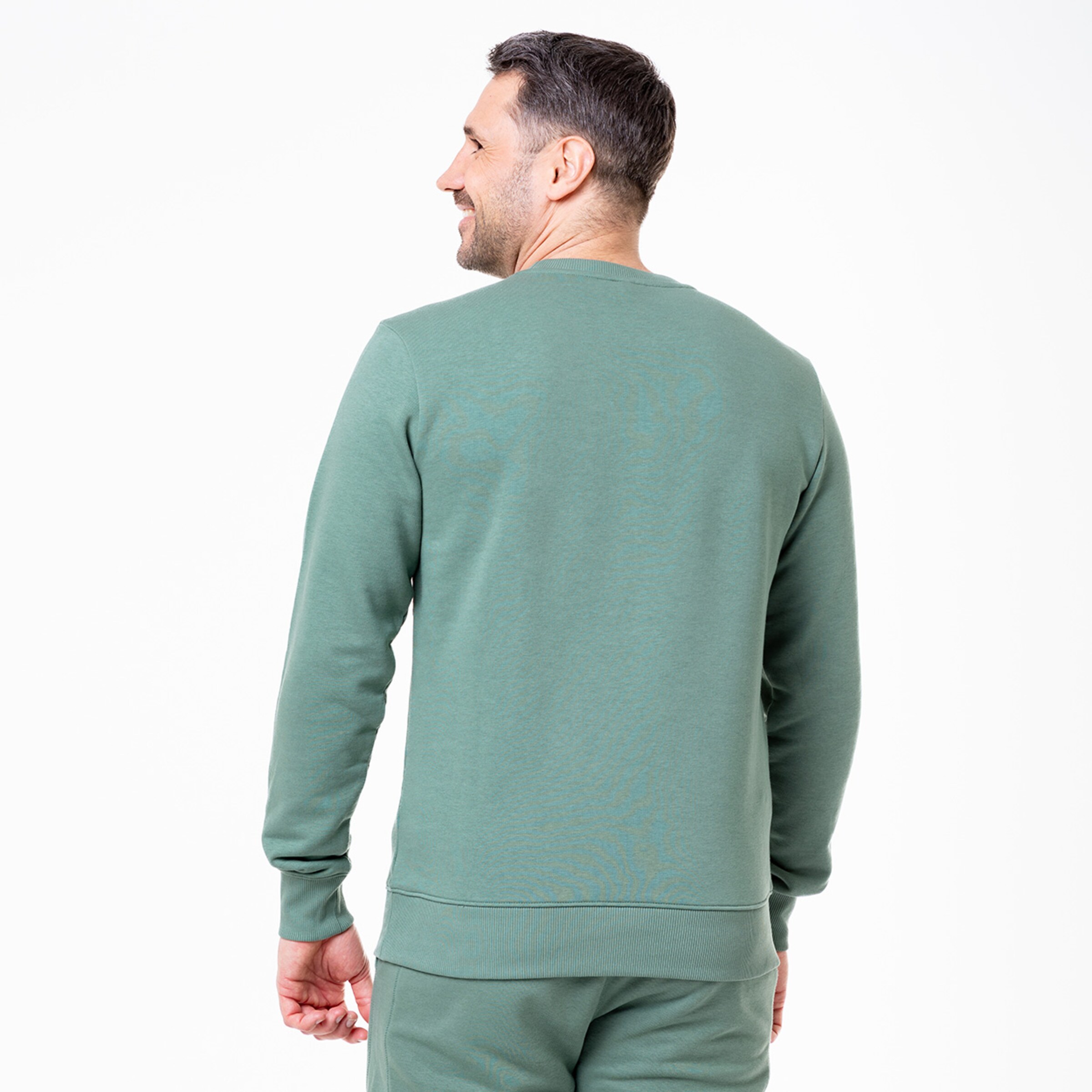 Mann mit kurzen Haaren trägt einen dunkelgrünen Pullover steht mit dem Rücken zur Kamera.