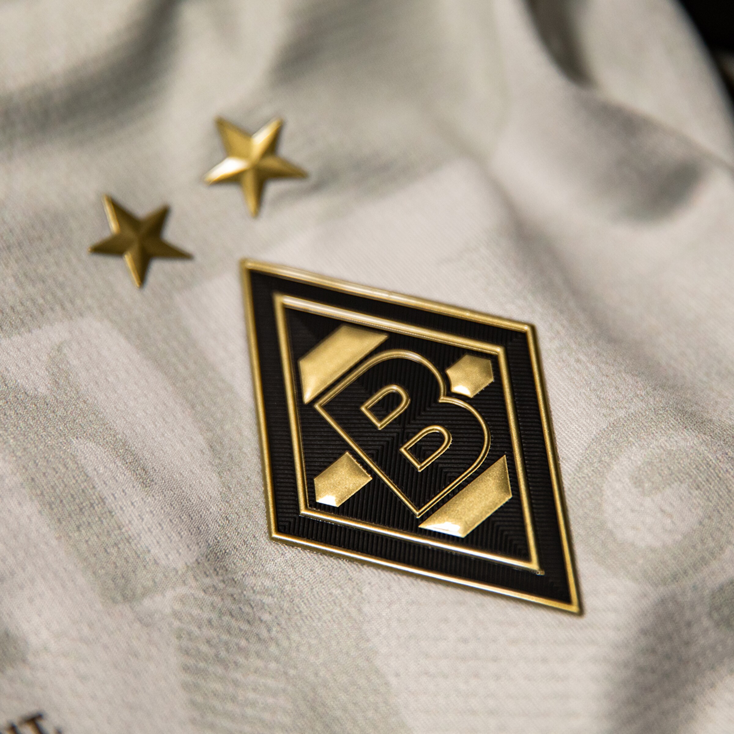 Nahaufnahme des gold-schwarzen Borussia Mönchengladbach Rauten-Logos mit zwei Sternen auf hellgrauem, strukturiertem Trikotstoff.