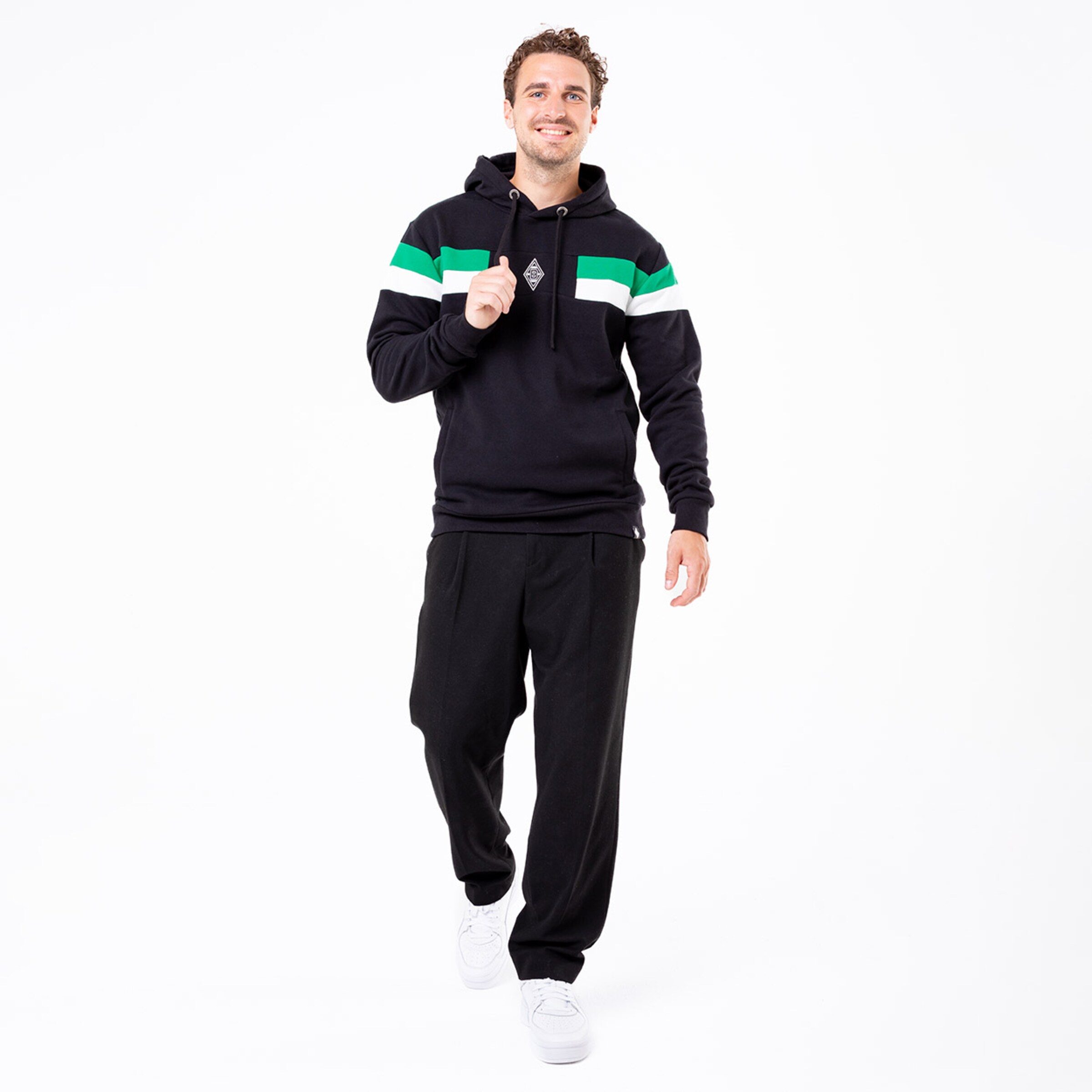 Ein Mann lächelt in einem schwarzen Hoodie mit grünen Streifen und schwarzen Jogginghosen. Er läuft aufrecht.