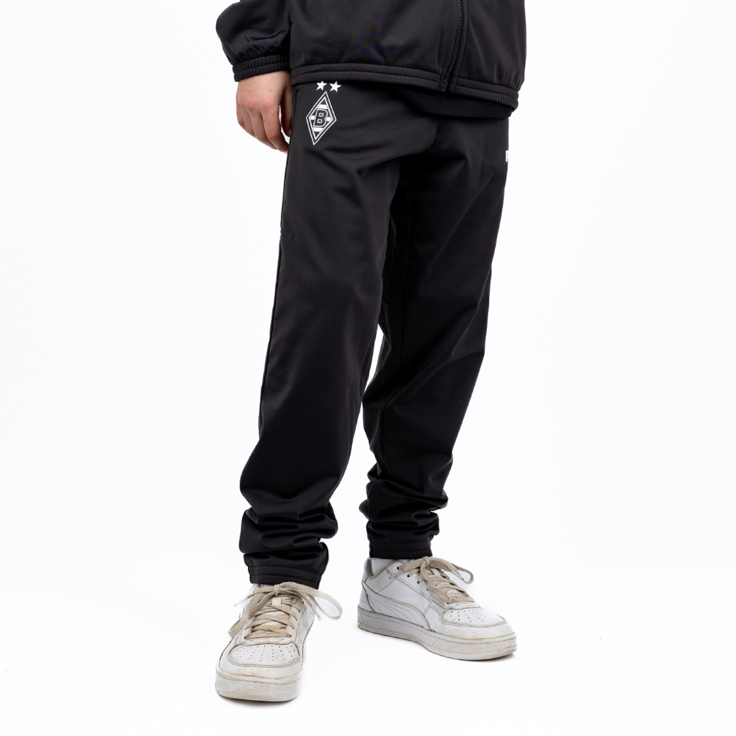 Person wearing black Borussia Mönchengladbach track pants with club logo and white sneakers against a white background.