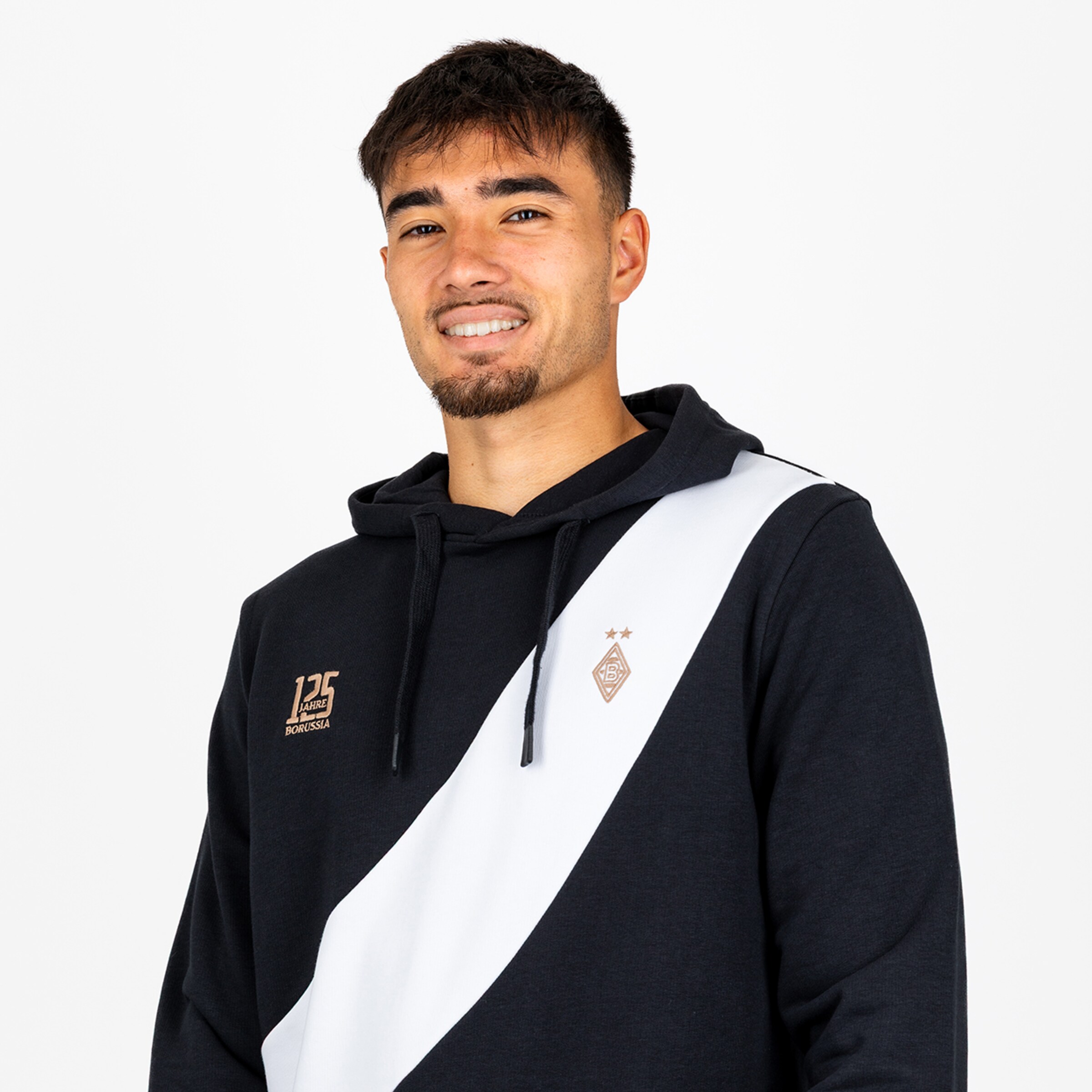 Porträt von Nathan Ngoumou im schwarzen Borussia-Hoodie mit weißem Schrägstreifen und Jubiläumslogo vor weißem Hintergrund.