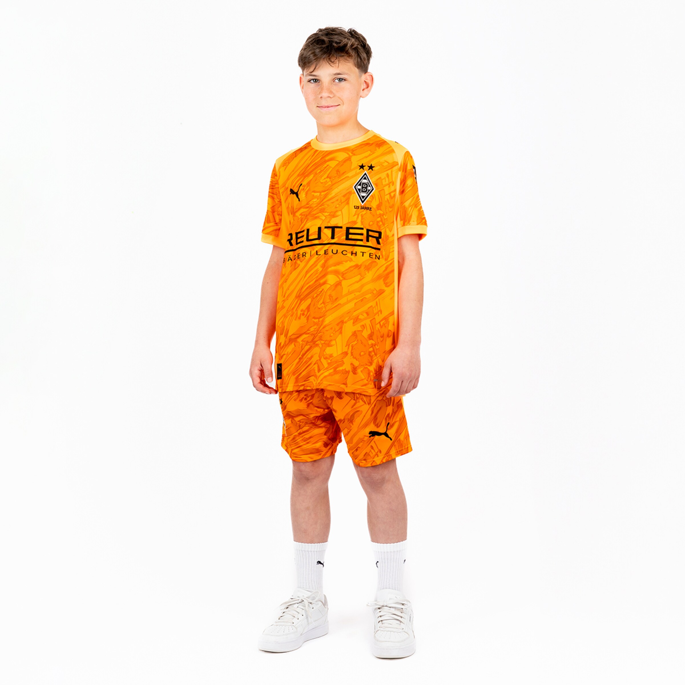Junge in orangefarbigem Borussia Mönchengladbach-Trikot und Shorts steht aufrecht mit einem neutralen Gesichtsausdruck.