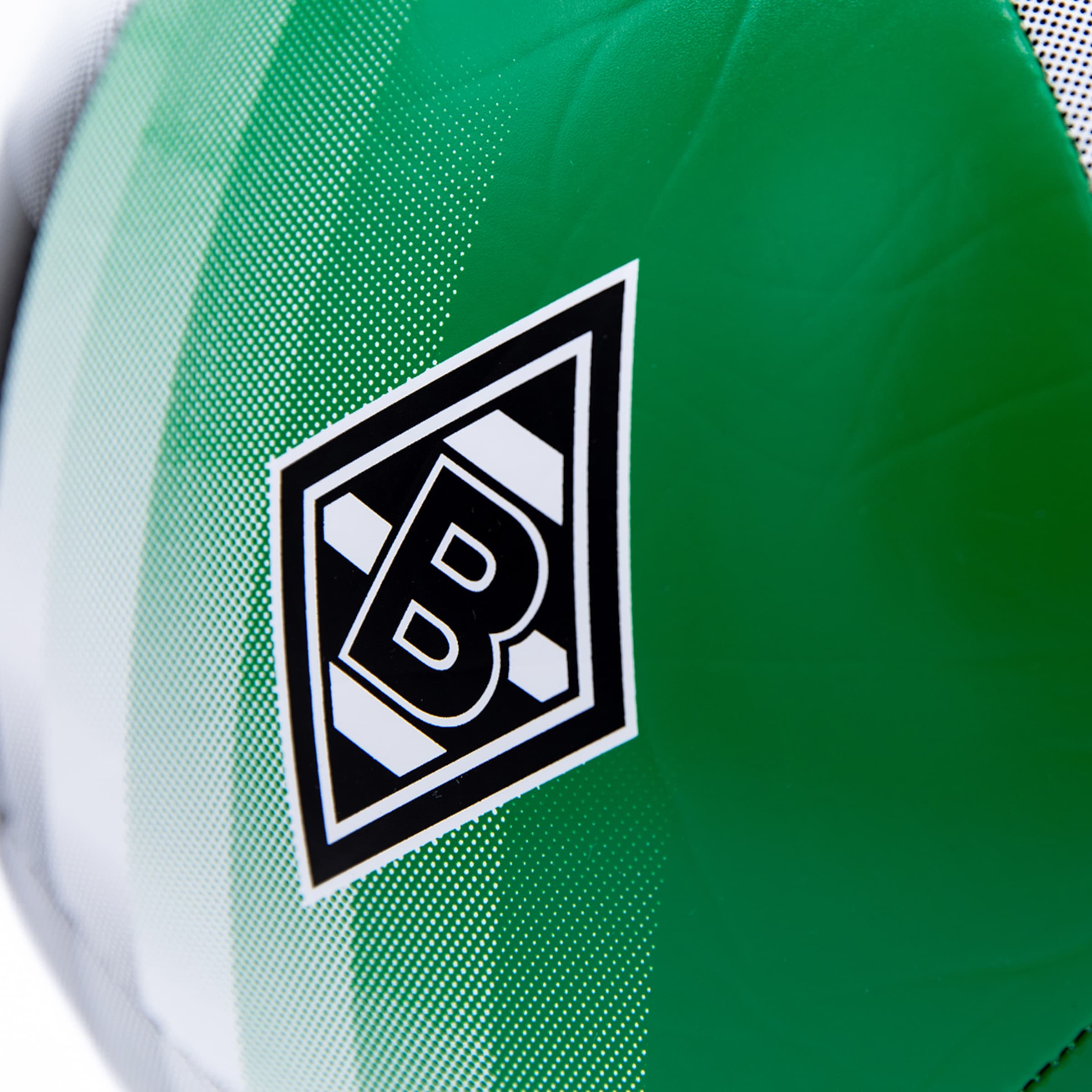 Nahaufnahme eines Fußballes mit dem Logo von Borussia Mönchengladbach auf einem grünen Feld mit gestreiften und gepunkteten Mustern. Der Ball hat eine glänzende Oberfläche.