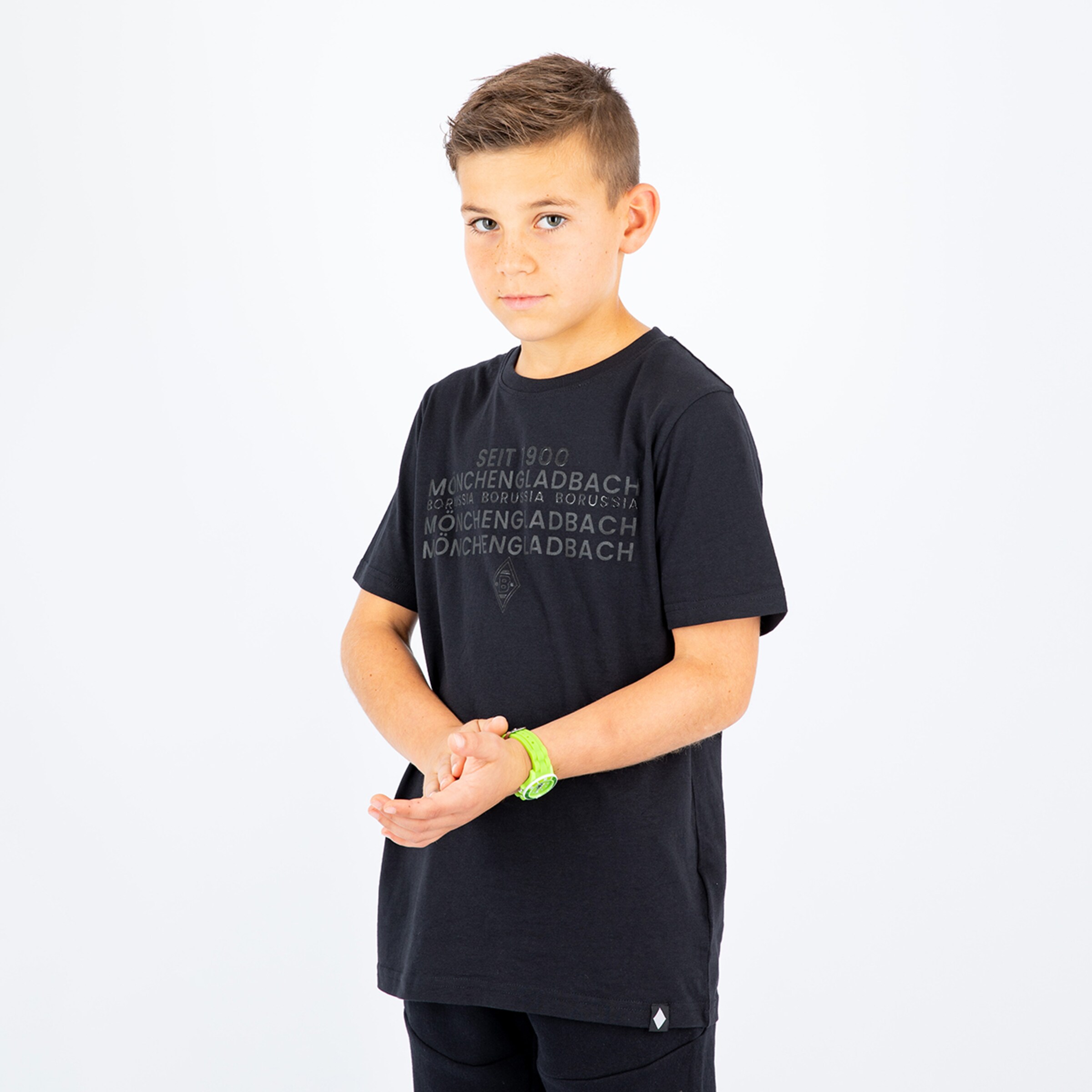 Ein Junge in einem schwarzen T-Shirt mit dem Text ‚Mönchengladbach‘ steht mit gefalteten Händen.