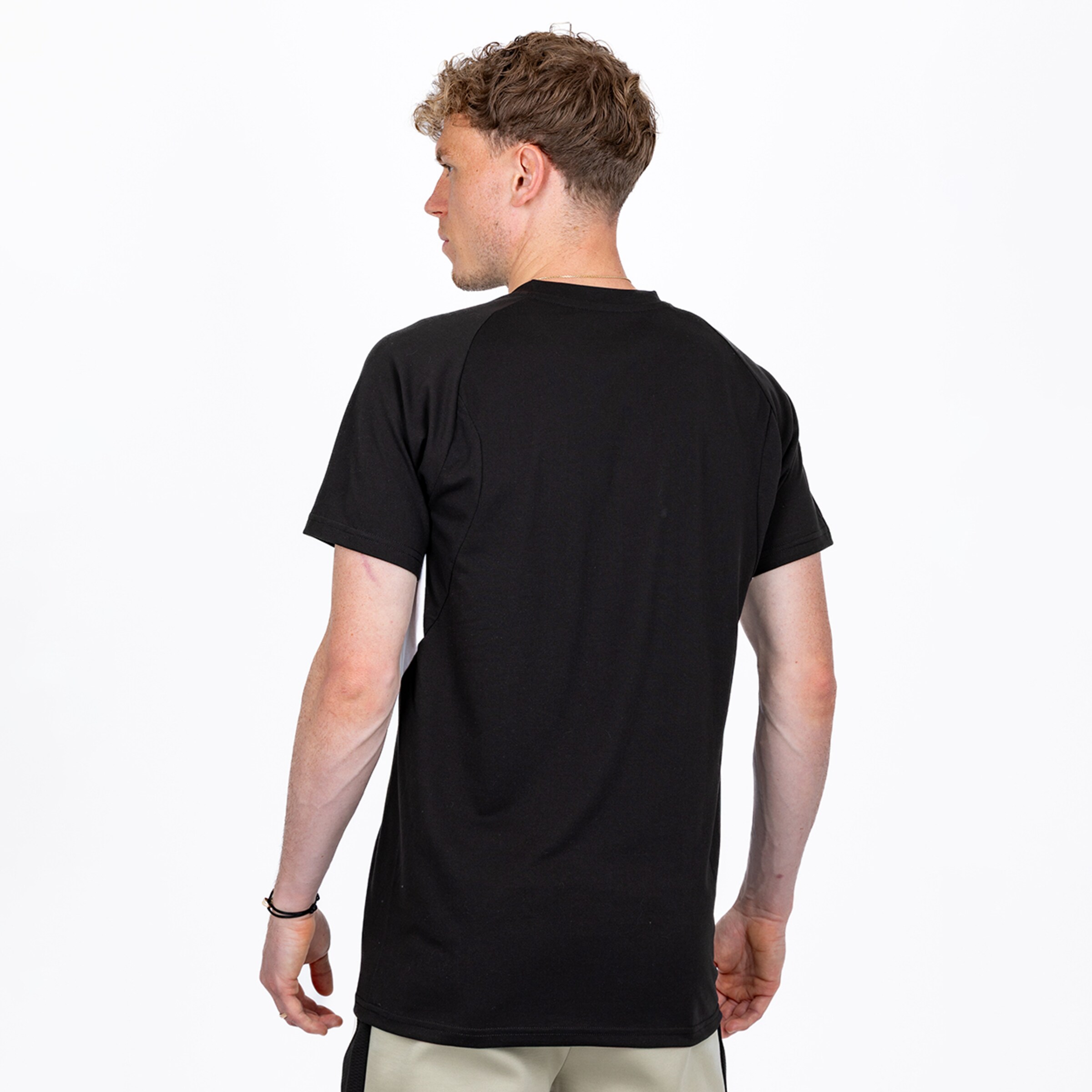 Rückansicht eines jungen Mannes mit lockigem braunem Haar, der ein schwarzes T-Shirt mit Raglanärmeln und hellen Shorts trägt und vor einem weißen Hintergrund steht.