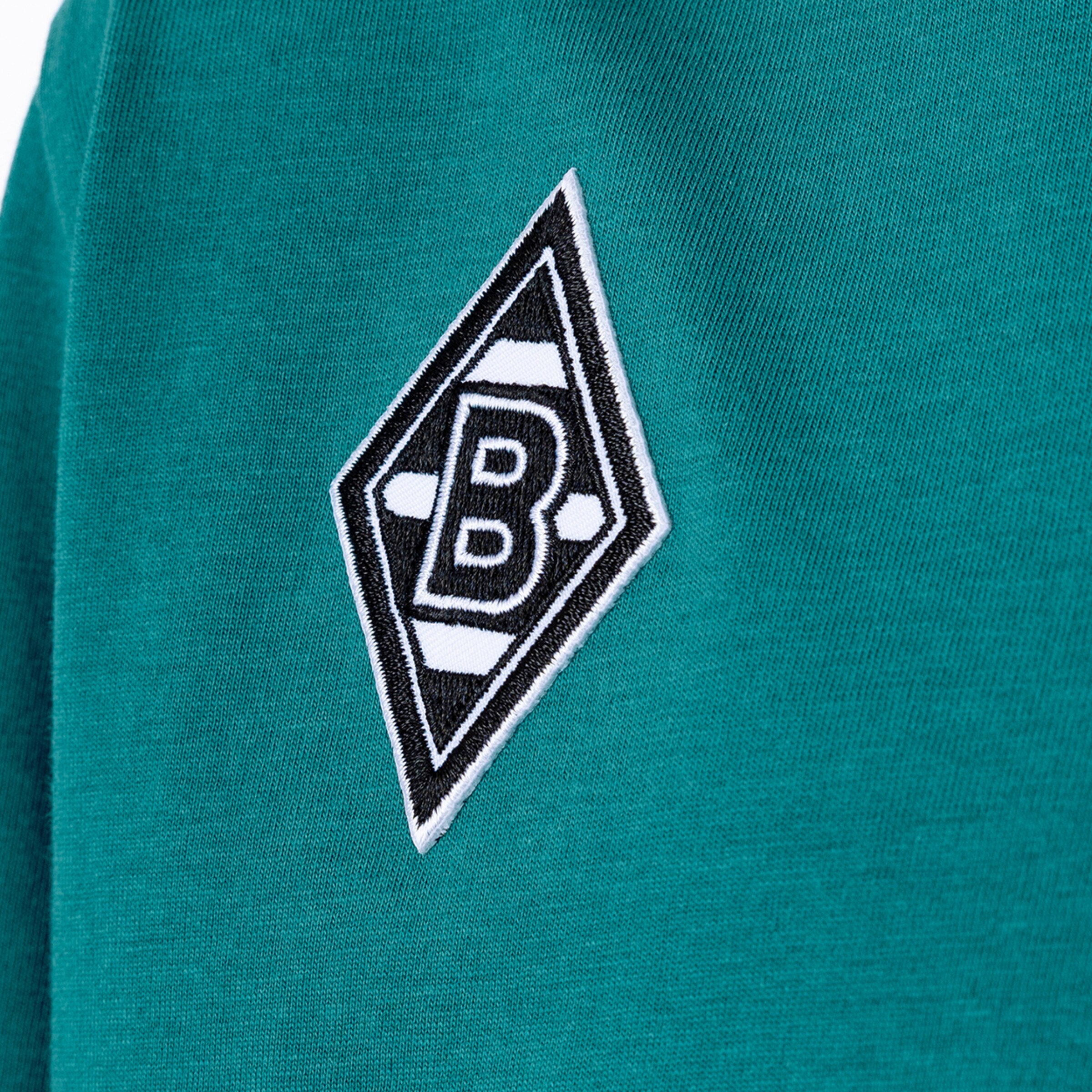 Close-up of the embroidered Borussia Mönchengladbach diamond logo with a black letter B, featured on teal-colored fabric.