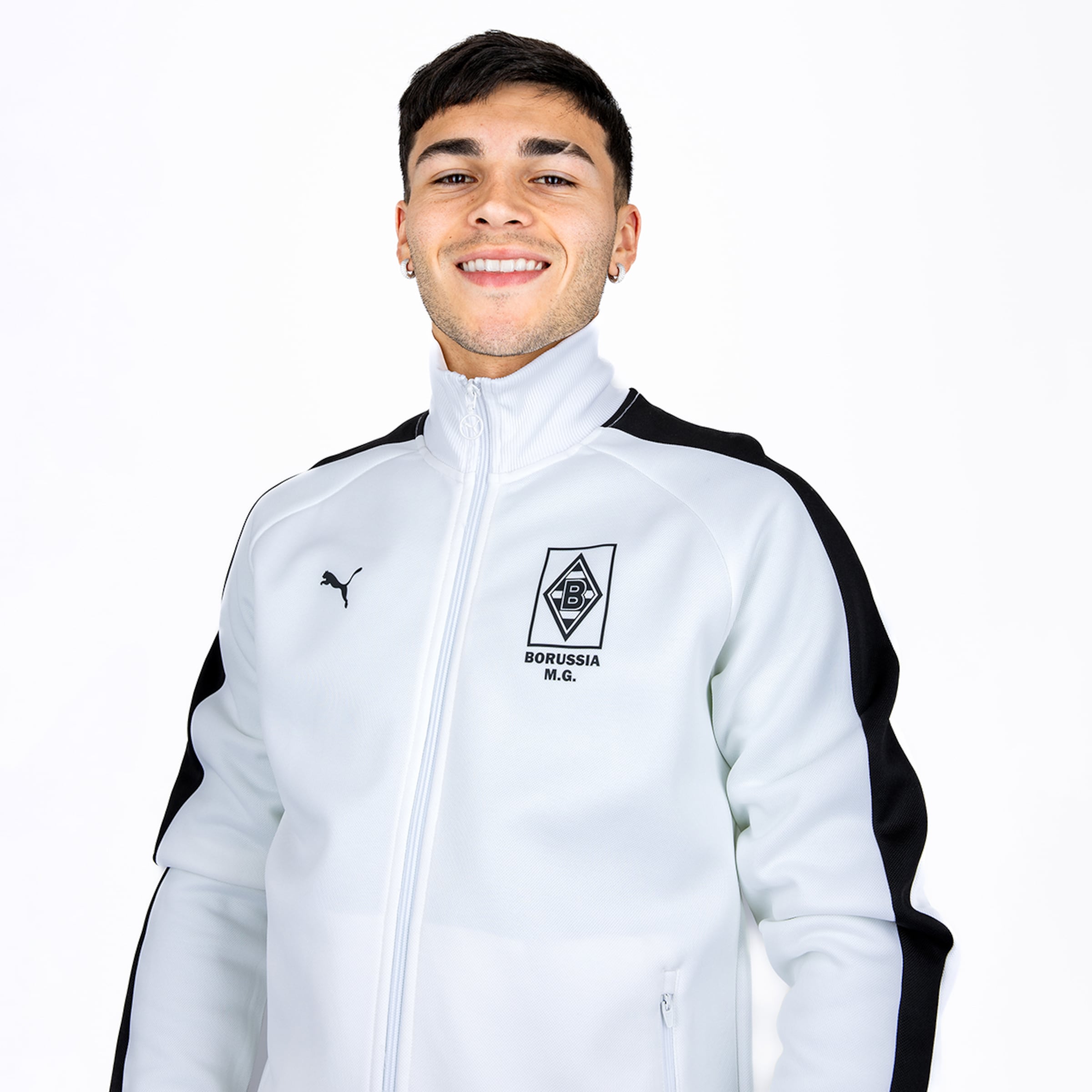 Junger Mann lächelt in weißer Puma-Trainingsjacke mit Borussia Mönchengladbach Logo vor weißem Hintergrund.